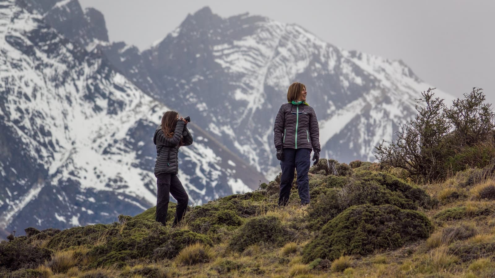 Two people, zero guide, and that wall of peaks entirely to themselves. Two women in down jackets stand on a mossy ridge during a self-guided W Trek in Patagonia, one photographing with binoculars raised while snow-capped granite peaks loom behind them under an overcast sky