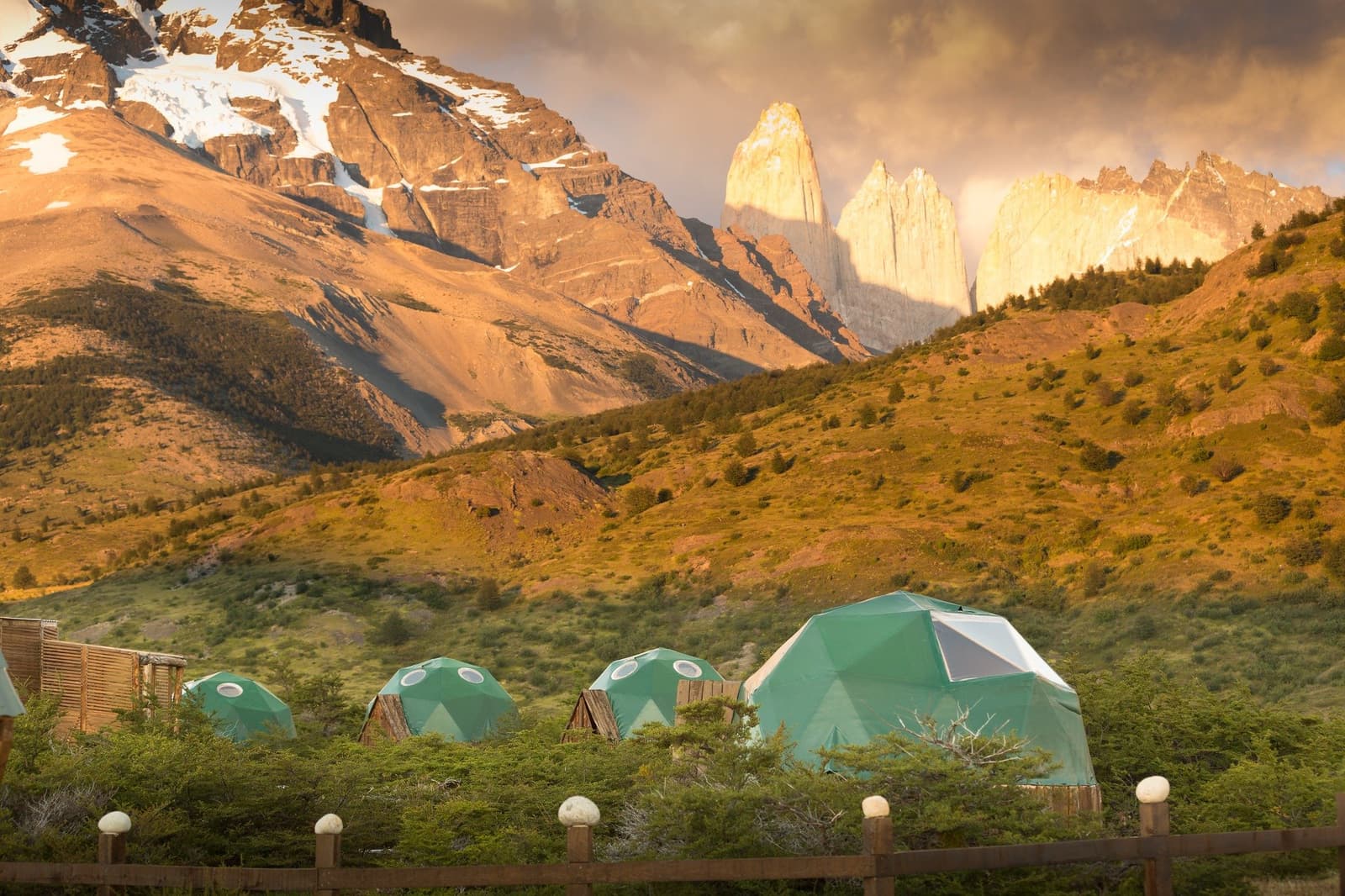 Your bedroom view comes pre-loaded with granite towers. Cluster of green geodesic domes at EcoCamp Patagonia during the short W trek, with the iconic granite towers of Torres del Paine glowing in warm evening light behind rolling hills