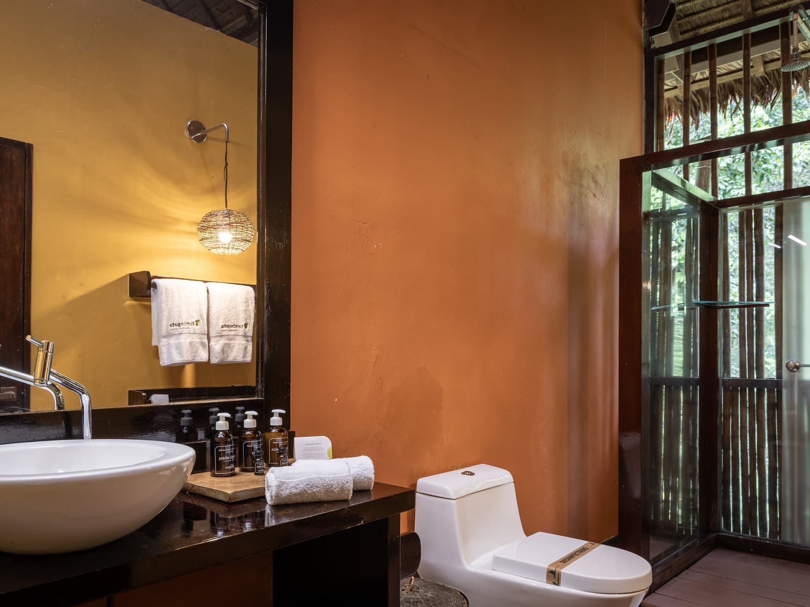 Deep in the Amazon, the forest outside your window, hot water inside. Private bathroom at the Tambopata Research Center Peru lodge, featuring a vessel sink, warm orange walls, wicker pendant light, branded toiletries, white towels, and a glass shower door with jungle foliage visible through the window