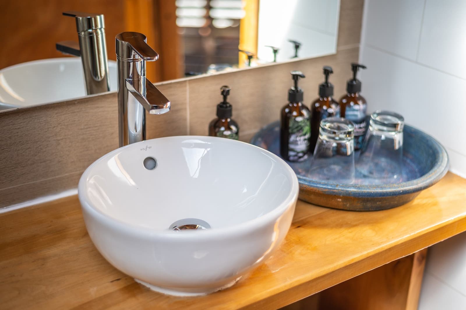 Refillable toiletries, real glassware, no single-use plastic in sight. Photo: Suite Dome Round vessel sink on a warm wooden vanity with a tall chrome faucet, beside a ceramic tray holding amber pump-bottle toiletries and glass tumblers, inside an eco-dome bathroom on the torres del paine 4 day all inclusive tour