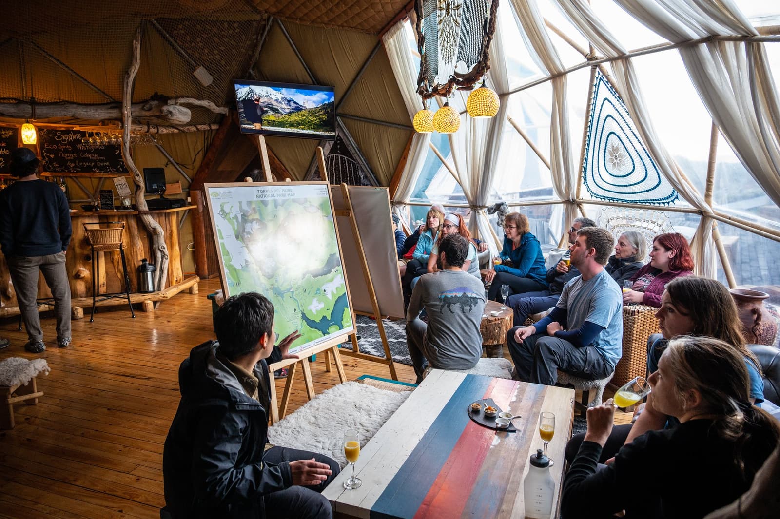 The map goes up, the questions start, and suddenly the W Trek feels very real. A guide presents a Torres del Paine W Trek map on an easel to a group of travellers seated inside a geodesic dome lodge, with drinks in hand and a TV screen showing park scenery in the background