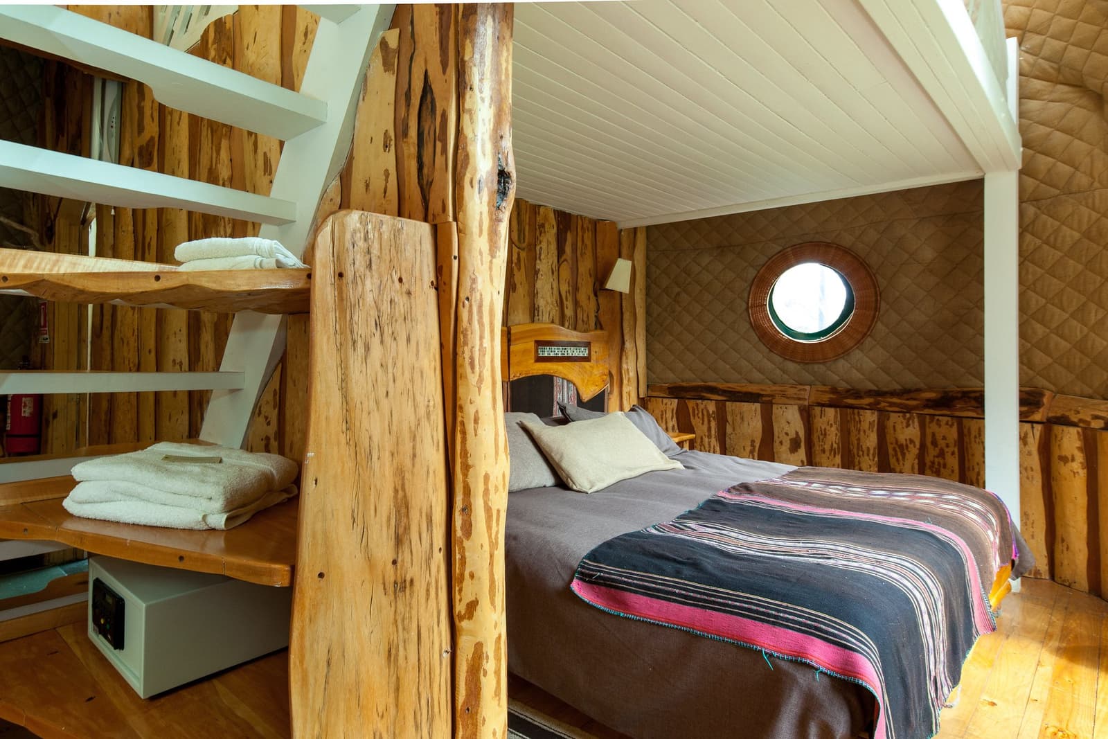 Madeira crua, manta colorida e uma janelinha redonda, o suficiente pra dormir bem. Photo: Suite Dome Loft Interior rústico de quarto em domo durante o trek W de Torres del Paine, com cama de casal coberta por manta listrada de tear, paredes de madeira bruta, janela circular tipo vigia e escada de madeira com toalhas dobradas