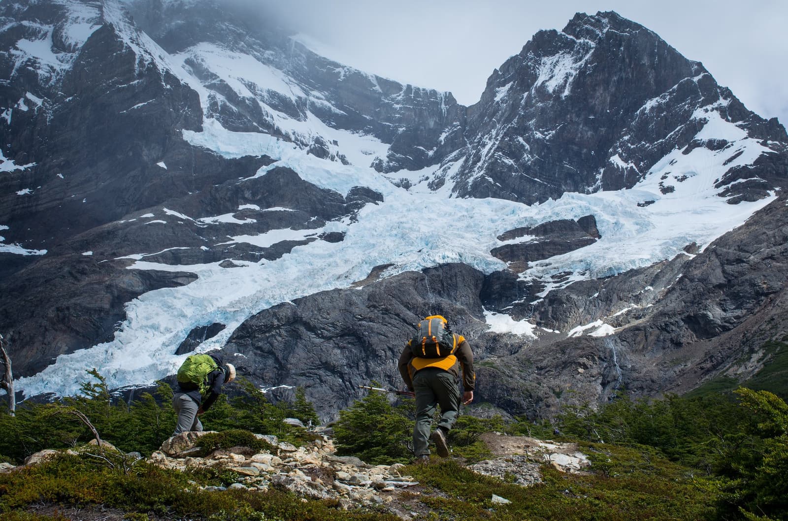 The glacier isn't getting any closer until you put one foot in front of the other. Two hikers with backpacks climbing a rocky trail on the torres del paine w trek, with a blue glacier spilling down a dark granite mountain face directly ahead