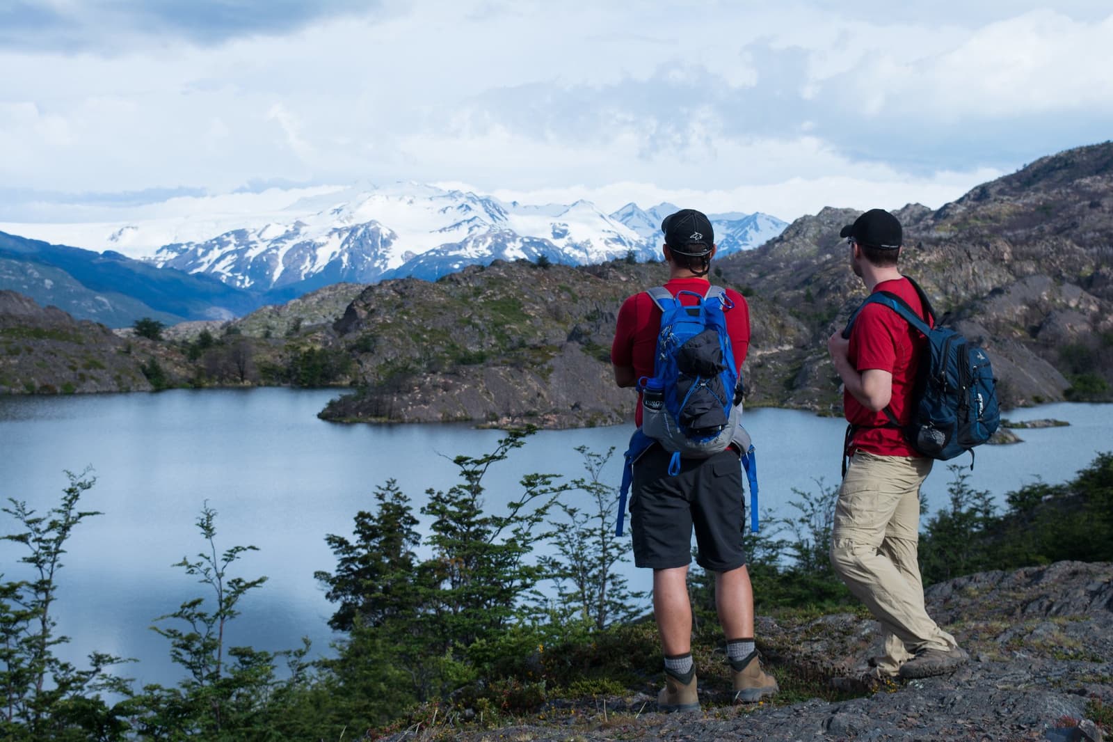 Two pairs of boots, one very good reason to stop and take it all in. Two hikers with backpacks stand on a rocky ridge on the Torres del Paine W Trek, looking out over a calm glacial lake surrounded by rugged hills and snow-covered mountains in the distance