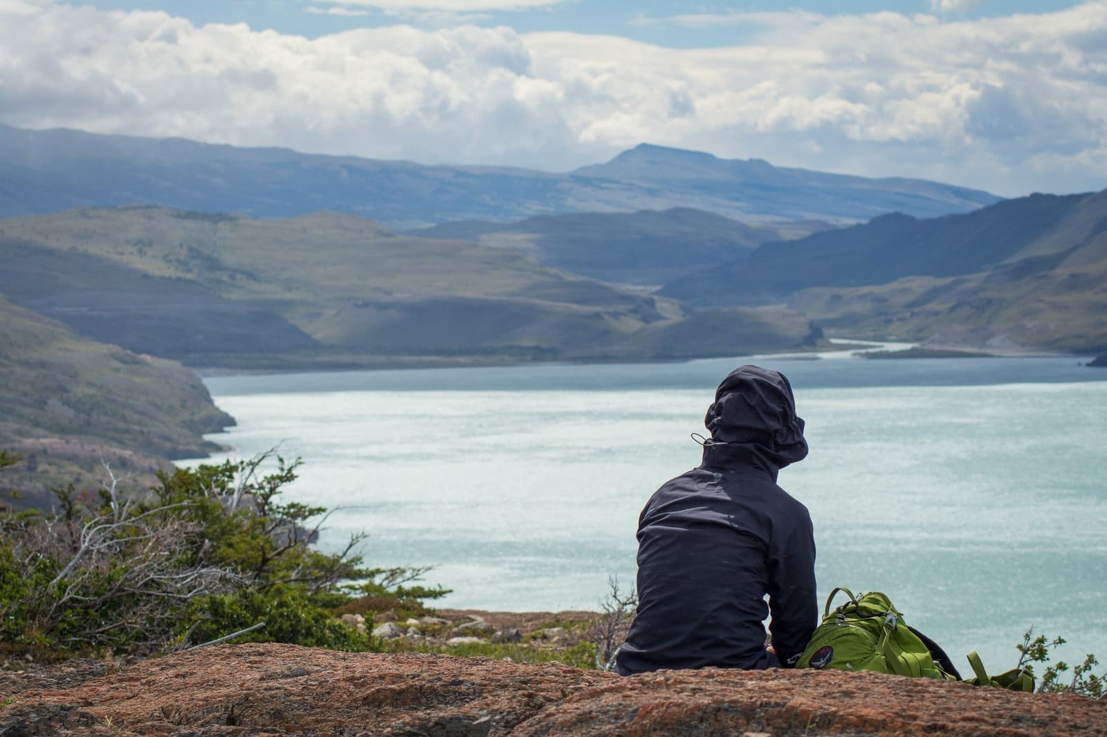 Lunch break with a view that makes you forget you earned it. A hiker in a dark jacket sits on a granite outcrop with a green backpack beside them, looking out over a turquoise glacial lake flanked by rolling green hills and distant mountains on the torres del paine w trek