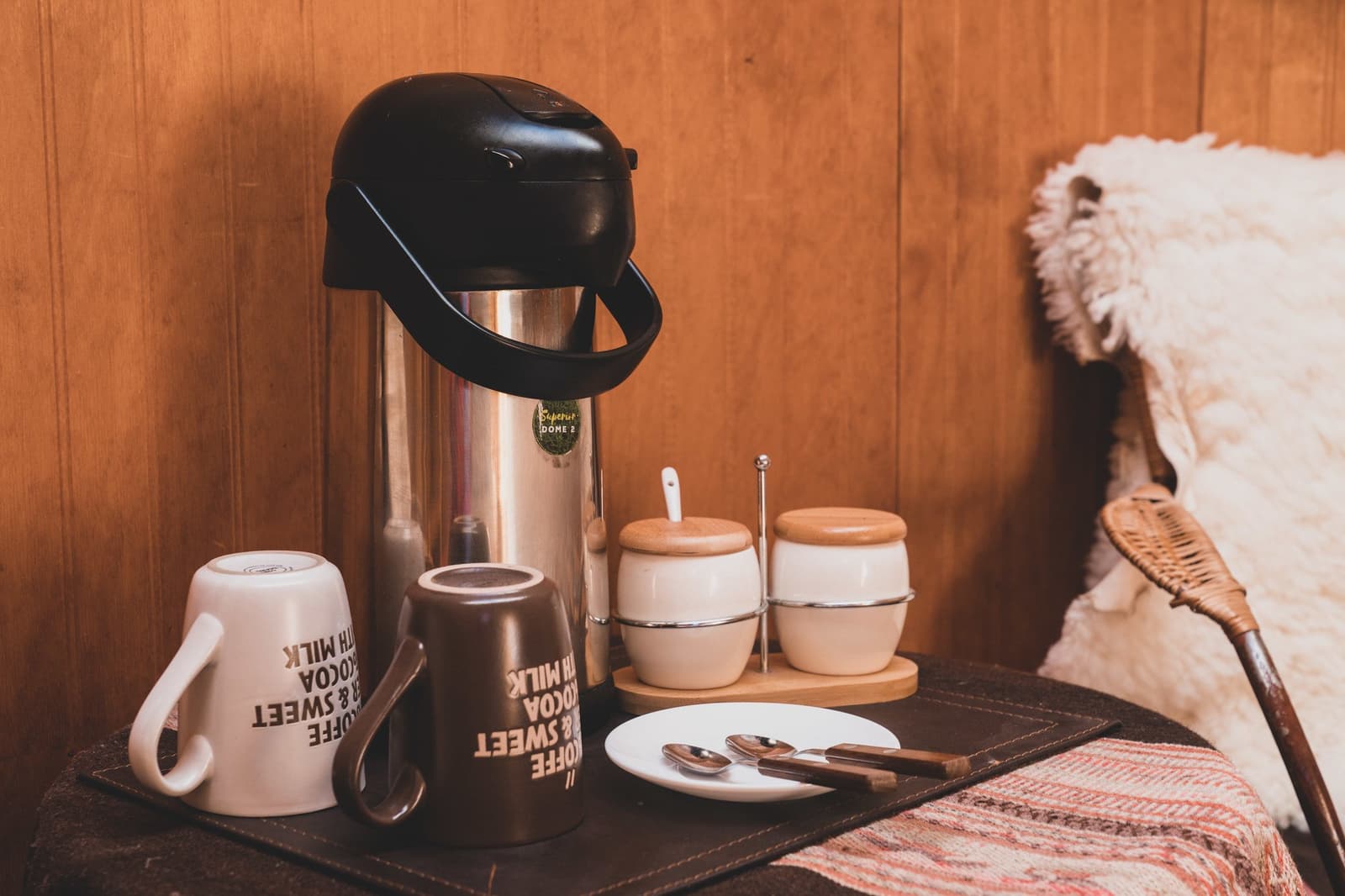 Café pronto antes de você amarrar as botas. Photo: Superior Dome Uma garrafa térmica de aço inoxidável com bomba, duas canecas com inscrições de café e cacau, potinhos de cerâmica para açúcar e creme em uma bandeja de madeira, ao lado de uma manta de pele de ovelha e uma cadeira de vime junto a uma parede de madeira em um domo EcoCamp no W Trek de Torres del Paine