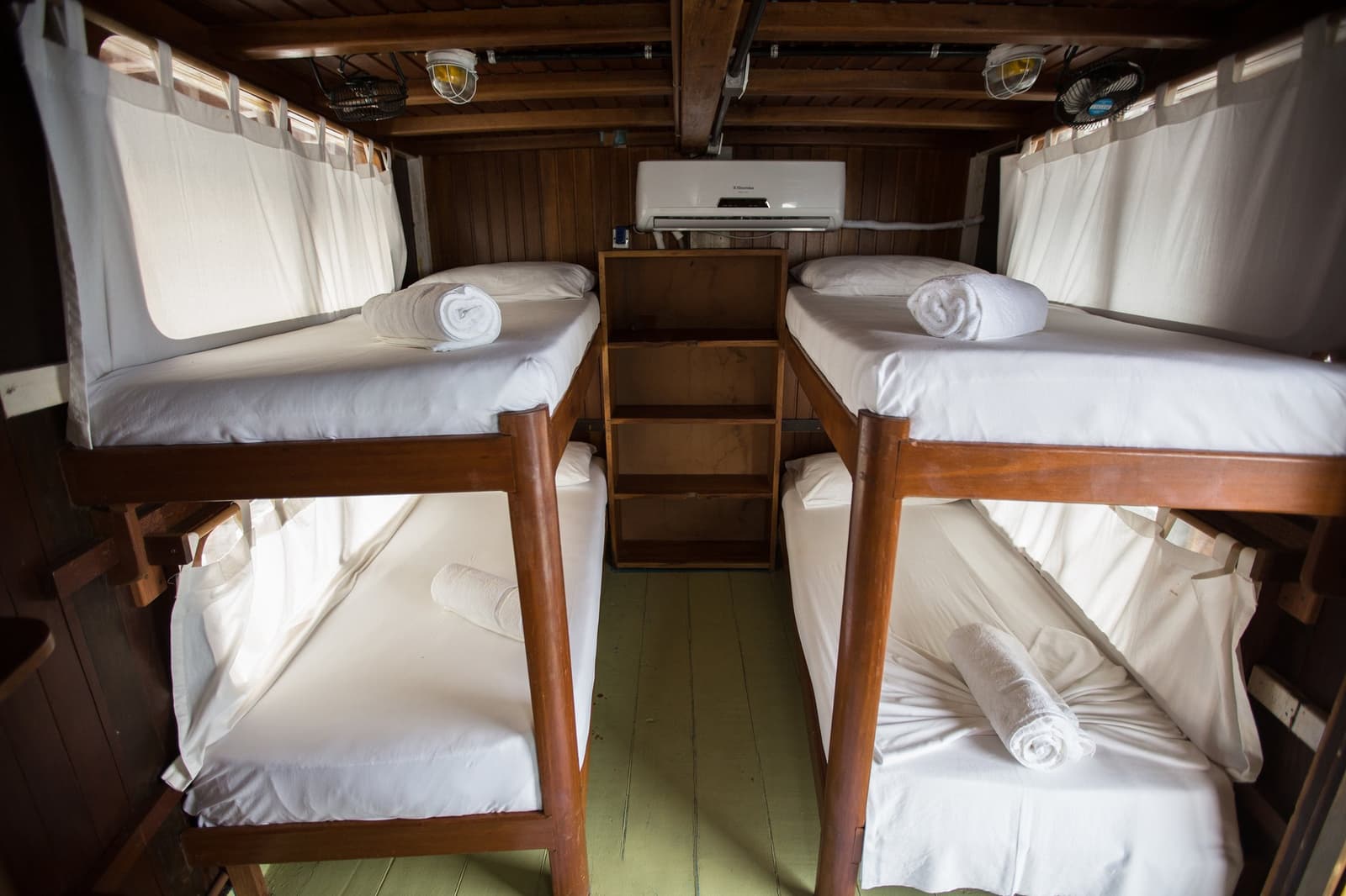 Four bunks, fresh linens, and a river outside every porthole. Photo: sitah Interior of a wooden-panelled cabin on an amazon cruise trip in Brazil, with two sets of bunk beds dressed in white linens, rolled towels on each mattress, white curtains, a wall-mounted air conditioner, and a central wooden shelf unit