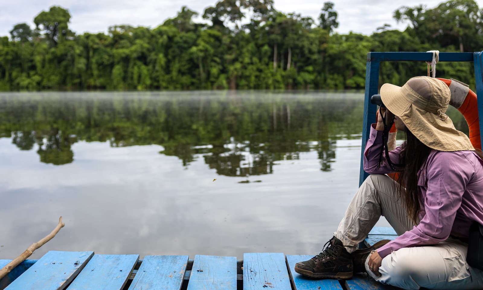 Binoculars, flat water, and zero distractions. This is your office for the morning. A woman in a wide-brim sun hat sits on the deck of a wooden river boat, looking through binoculars across a glassy Amazon river toward dense rainforest on this amazon tour from puerto maldonado