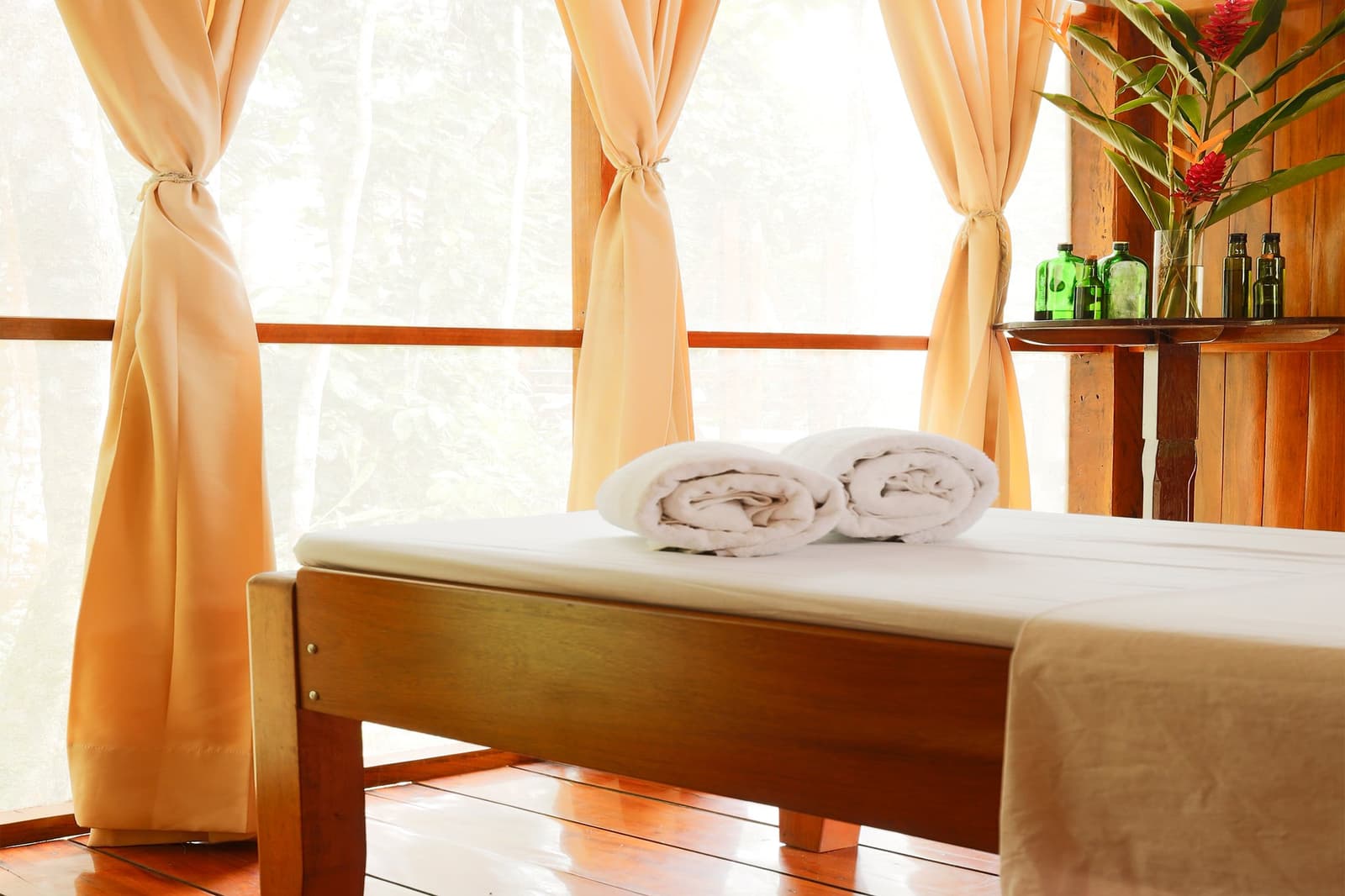 After a day on the river, the forest can wait a little longer. A wooden massage table with two rolled white towels sits in a warm spa room at Posada Amazonas, with sheer curtains filtering forest light through the windows and glass oil bottles on a shelf beside a red tropical flower, as part of an amazon tour from puerto maldonado