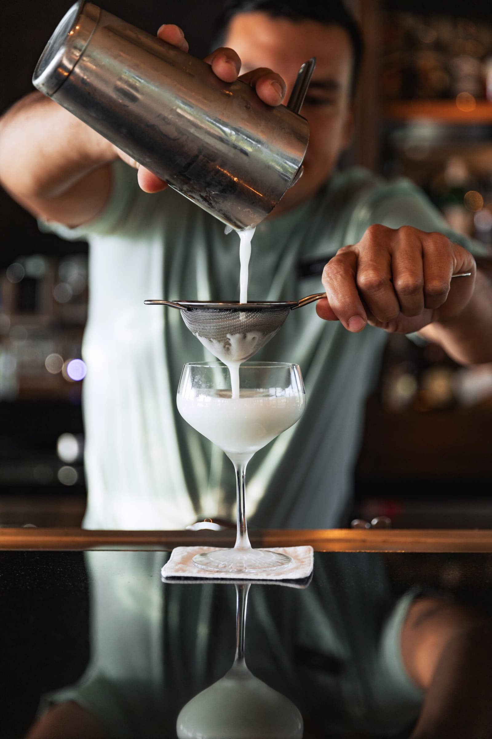 Fine straining, no shortcuts. Sundowners on the river deserve the full treatment. A bartender double-straining a frothy white cocktail through a fine mesh sieve into a coupe glass on the bar of the Aria Amazon cruise ship