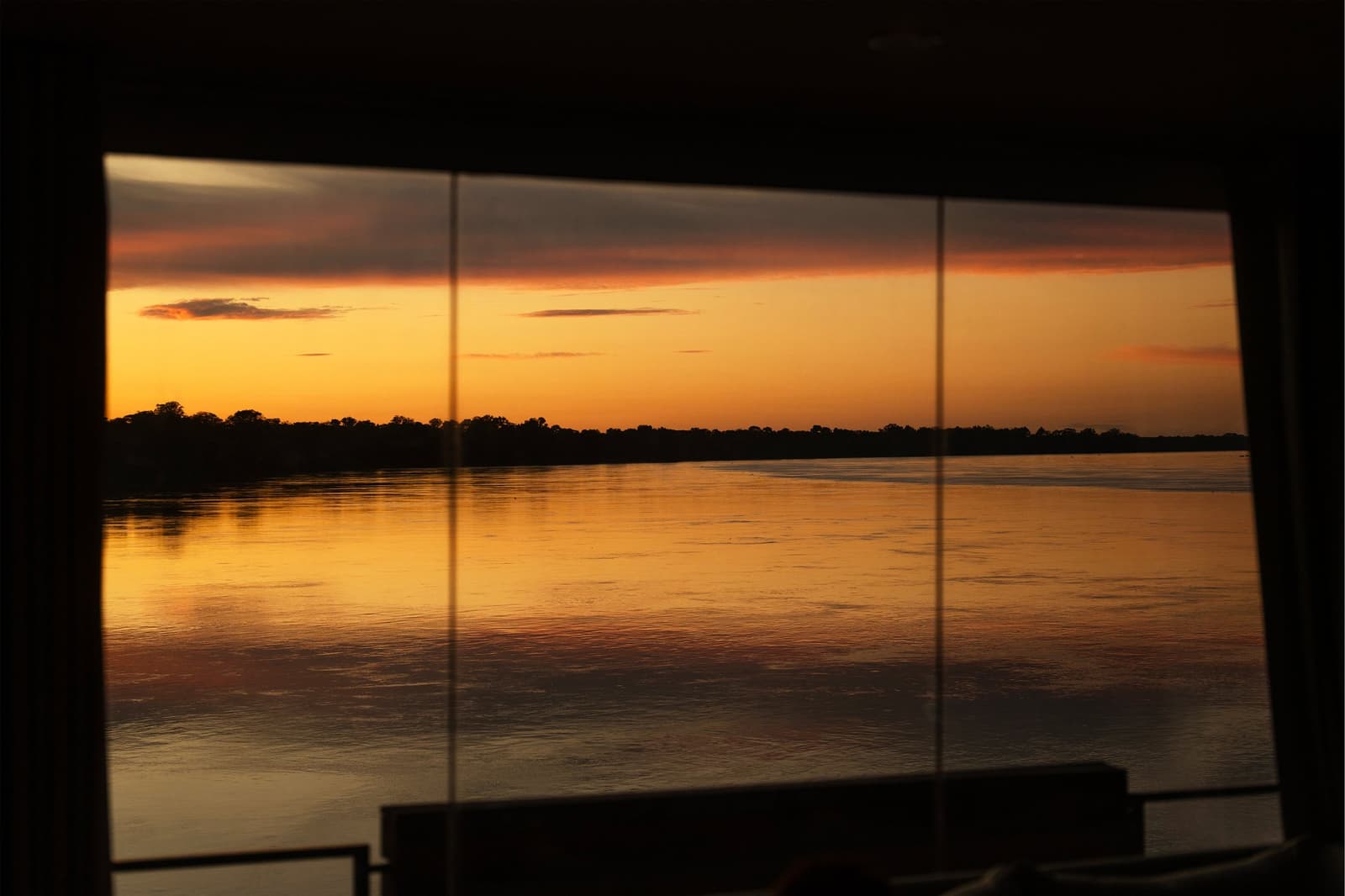 Some views earn a slow cup of coffee and nowhere to be. Photo: Daniel Silvia Yoshisato Golden sunset over a wide Amazonian river seen through the floor-to-ceiling panoramic windows of the Aria Amazon cruise ship, with a dark tree-line silhouette reflected on the calm water below an orange-tinted sky