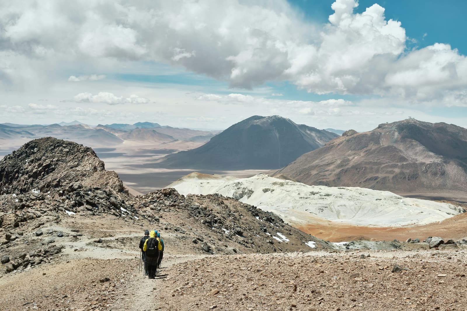 A esta altitud, el paisaje no te regala nada, todavía queda bajar. Dos senderistas con bastones de trekking y mochilas cargadas descienden una cresta rocosa de gran altitud en una expedición de camping en el desierto de Atacama, con un amplio paisaje volcánico, depósitos de azufre pálidos y cumbres cónicas oscuras extendiéndose hasta el horizonte bajo un cielo parcialmente nublado