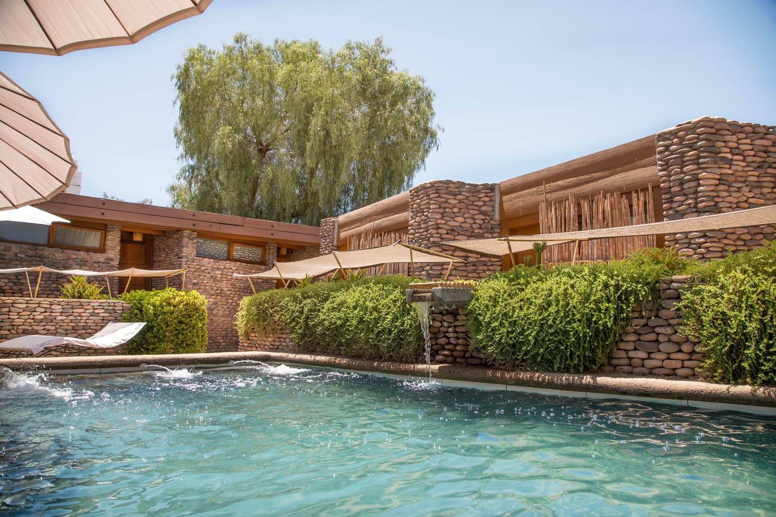 El chapuzón que te espera después de los senderos vale cada paso. Piscina exterior con cascada de piedra en un lodge del desierto, rodeada de muros de canto rodado, vallas de caña, velas de sombra y un gran sauce, como base refrescante entre las aventuras de camping en el desierto de Atacama