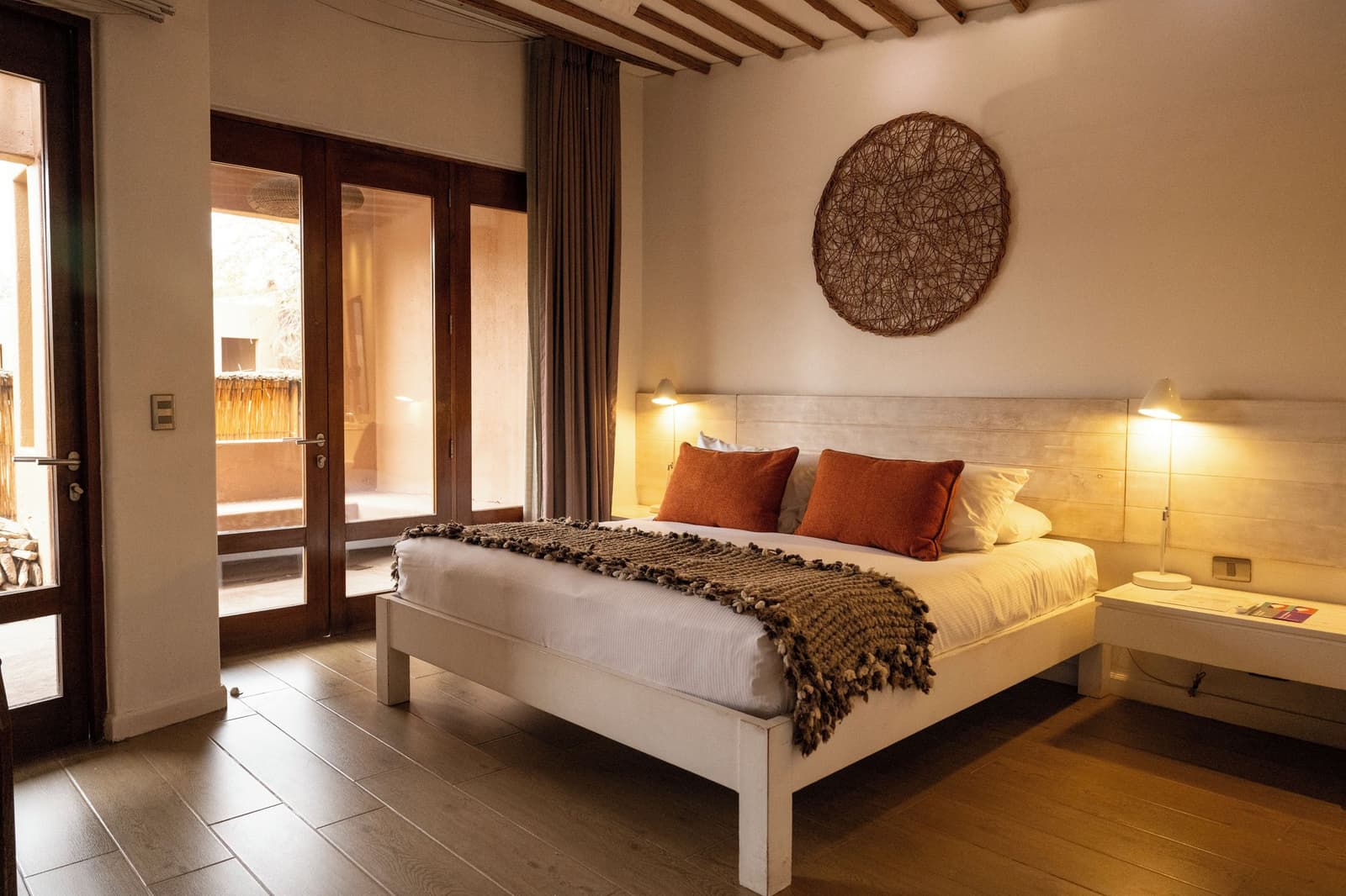 Terracotta cushions, warm lamp glow, and a courtyard just beyond the door. Warmly lit double bedroom in an atacama desert tour package lodge, featuring a white timber bed frame with terracotta cushions, a chunky knit throw, exposed wooden ceiling beams, a woven circular wall decoration, and French doors opening onto a sunlit courtyard
