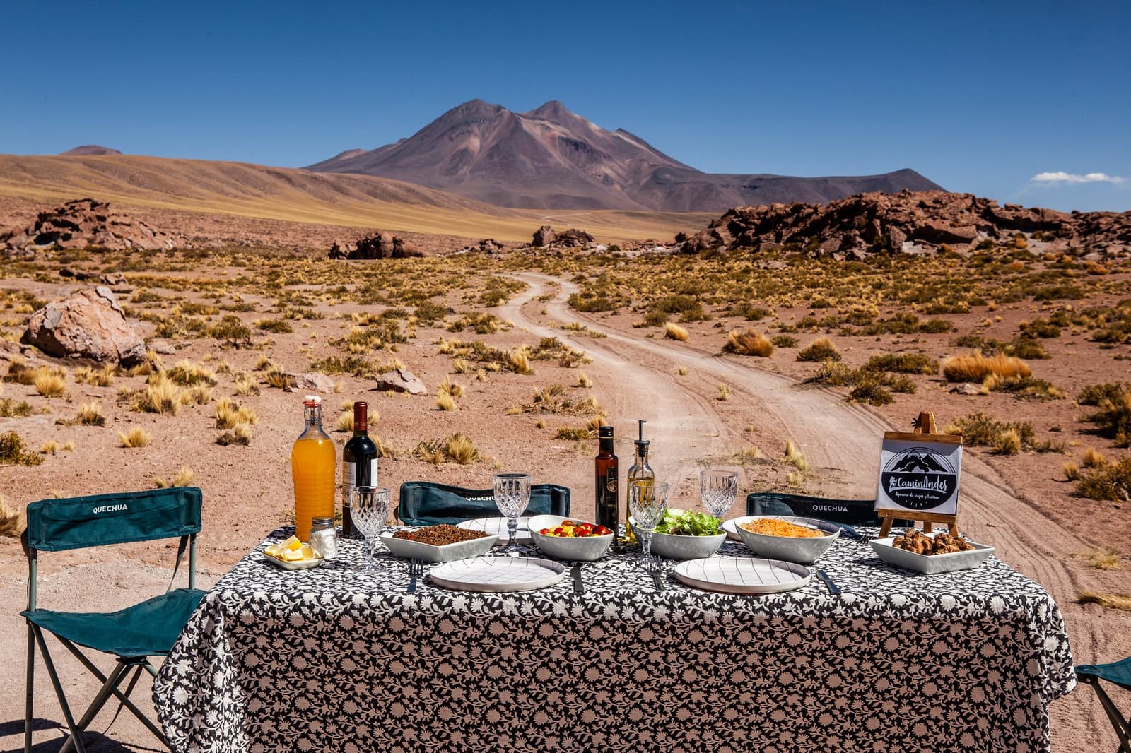 Lunch with a volcano for a backdrop. No reservation required. A fully set dining table with wine, juice, salads, and dishes laid out on a patterned tablecloth in the middle of the Atacama Desert, with a dirt track leading toward a volcanic peak under a deep blue sky — part of an atacama desert tour package experience