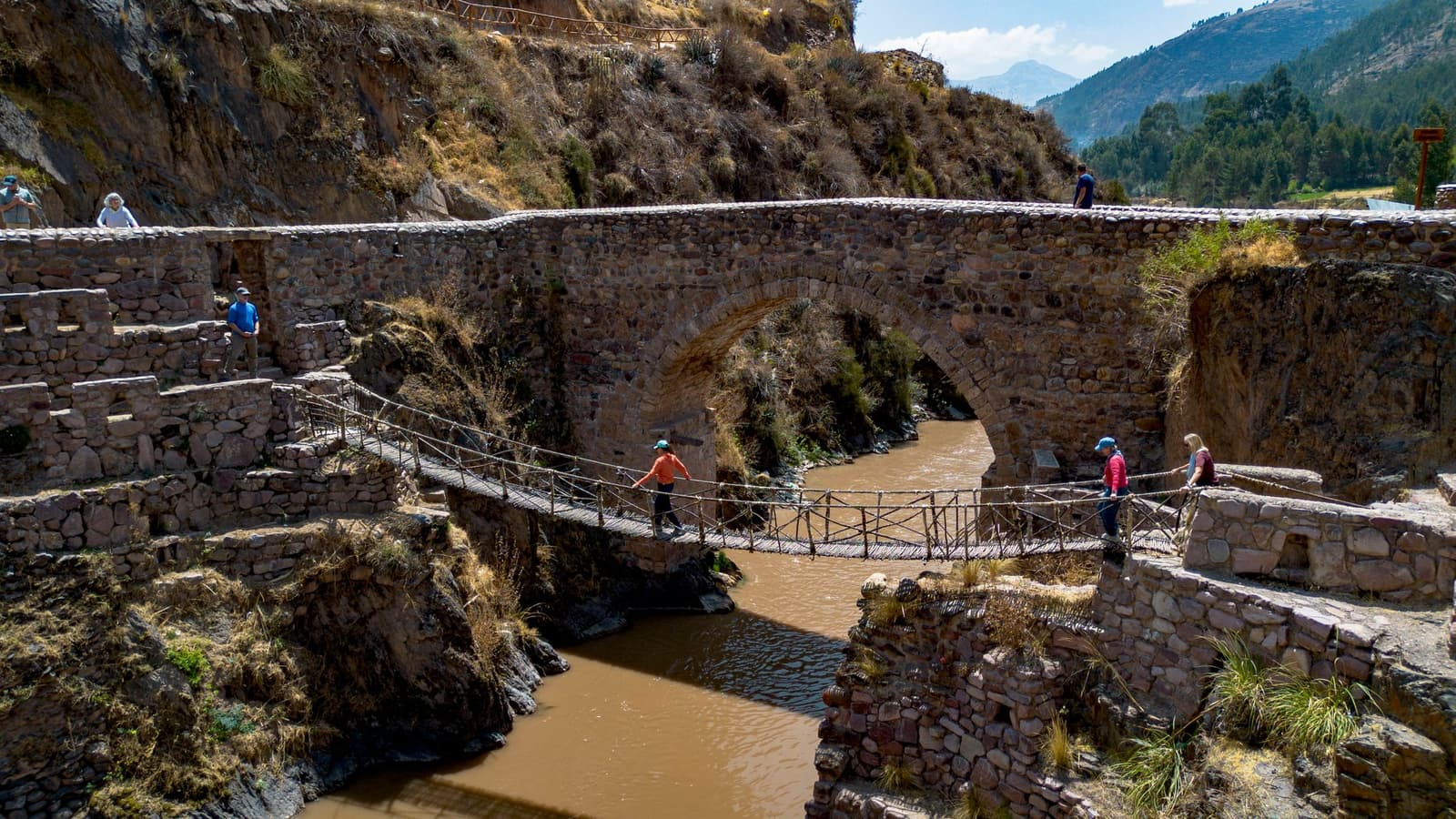 Two bridges, one river, five centuries between them, pick your crossing. Hikers crossing a narrow rope-and-plank suspension bridge over a muddy river, with a large colonial stone arch bridge rising behind them, set in a steep Andean canyon on the Ausangate Lodge Trek