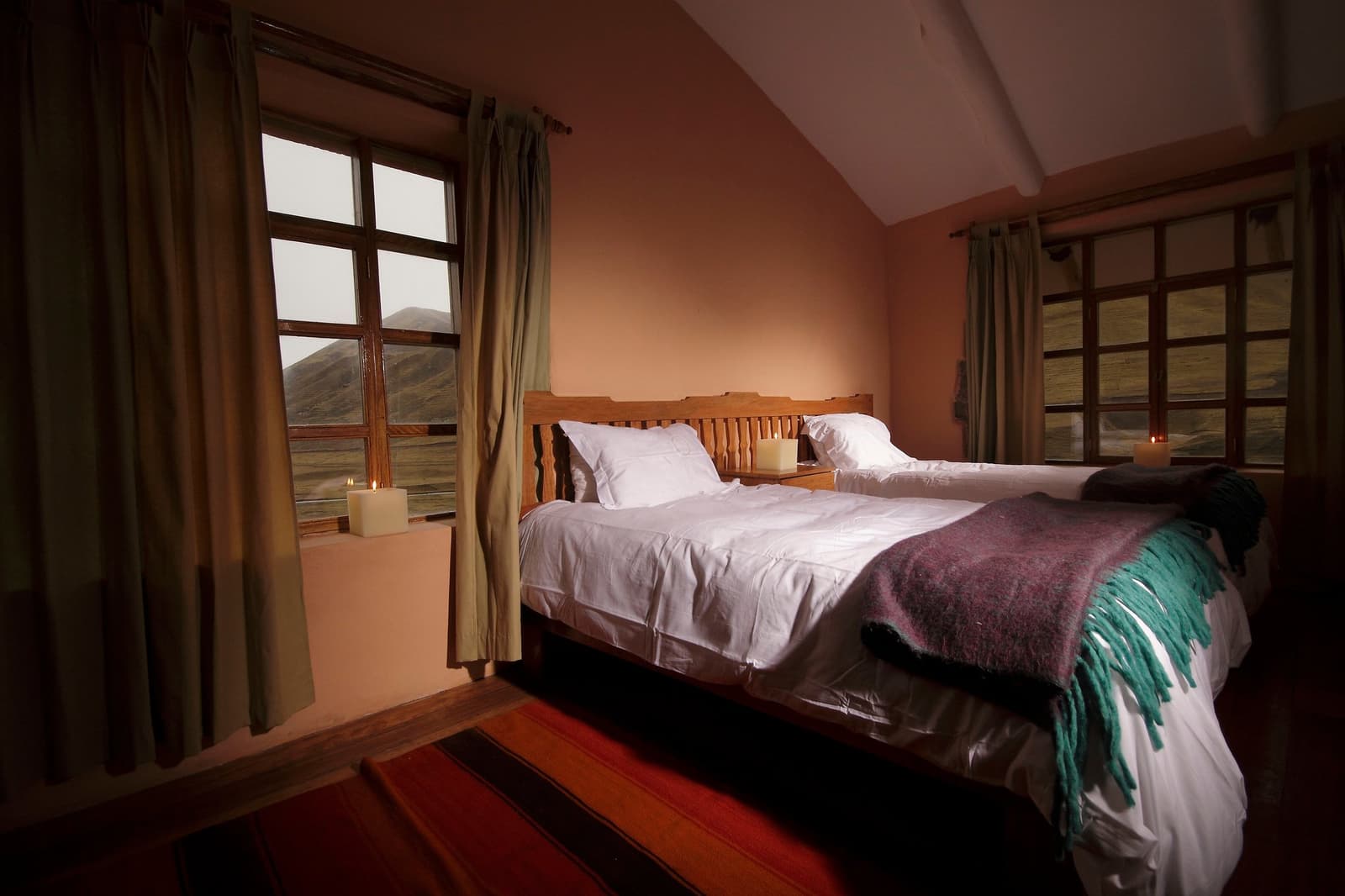 Candles, a wool throw, and hills right outside the window. Altitude has never felt this easy to come home to. Photo: Gustavo Vivanco Leon A double bed with white linen and a purple wool throw, lit by candles on wooden nightstands, with windows overlooking high-altitude Andean hills — part of the Ausangate Lodge Trek accommodation