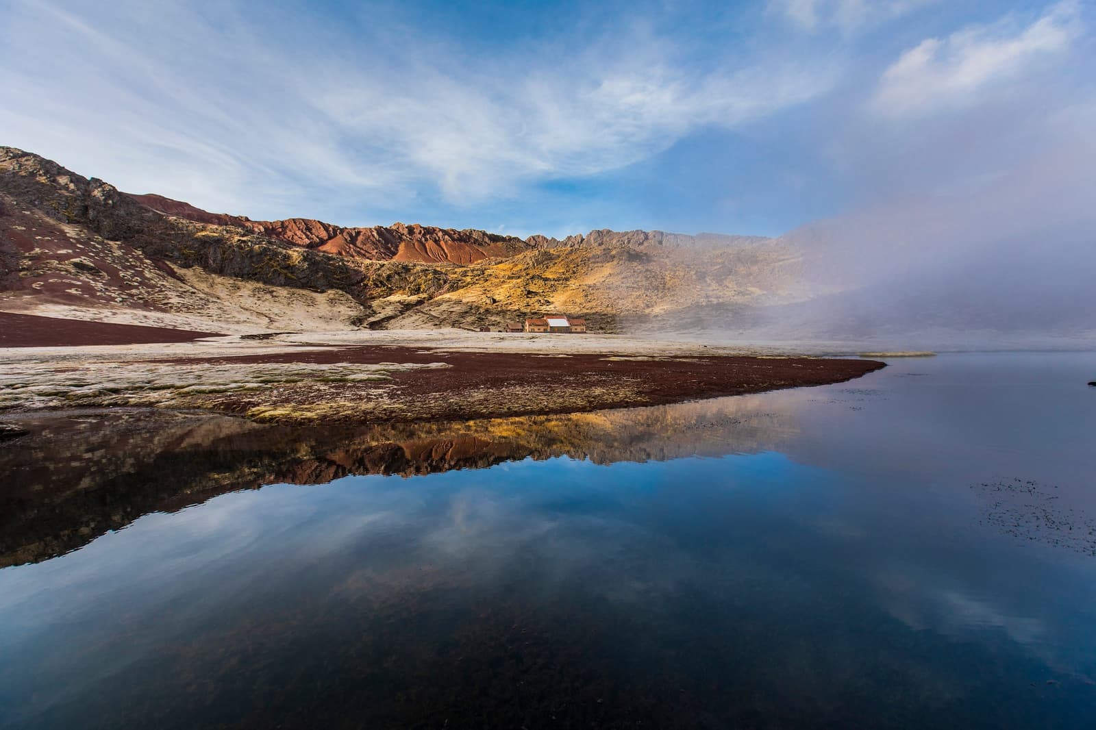 That tiny shelter across the water is your whole world for the next few hours. Photo: Paul Spierenburg A still high-altitude lake mirrors rugged red and ochre mountain slopes under a blue sky with low mist on the Ausangate Lodge Trek, with two small stone buildings visible on the far shore