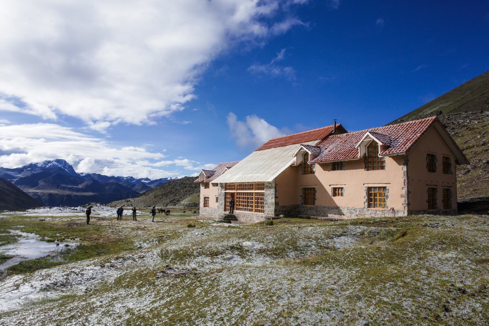Frost on the grass, a warm bed inside, and llamas somewhere nearby. A two-storey stone and plaster lodge with a terracotta-tiled roof sits on a frost-dusted high-altitude plateau during the Ausangate Lodge Trek, with snow-capped Andean peaks rising under a vivid blue sky and several trekkers standing outside