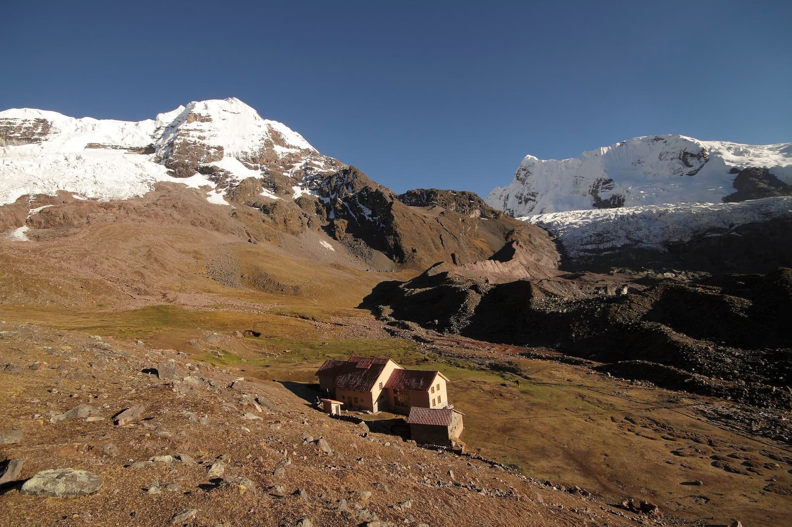 Tiny against the glaciers, the lodge still manages to feel like enough. Photo: Gustavo Vivanco Leon A small stone mountain lodge with a red-tiled roof sits on a rocky high-altitude slope during the Ausangate Lodge Trek, with two large snow-capped peaks rising sharply under a deep blue sky