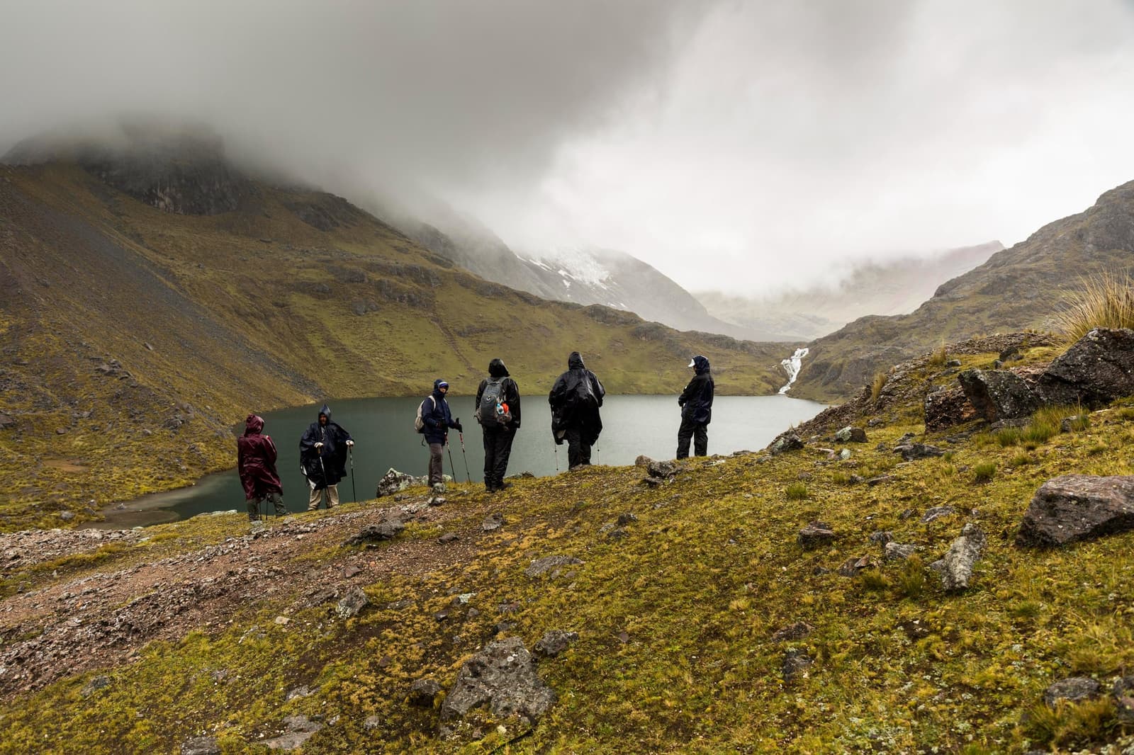 Rain on the ridge, and nobody's turning back. A group of six hikers in rain gear and ponchos pauses on a mossy, rocky ridge during the ausangate trek, looking out over a calm glacial lake cupped between steep green-and-grey Andean slopes, with snow-streaked peaks and low clouds in the background