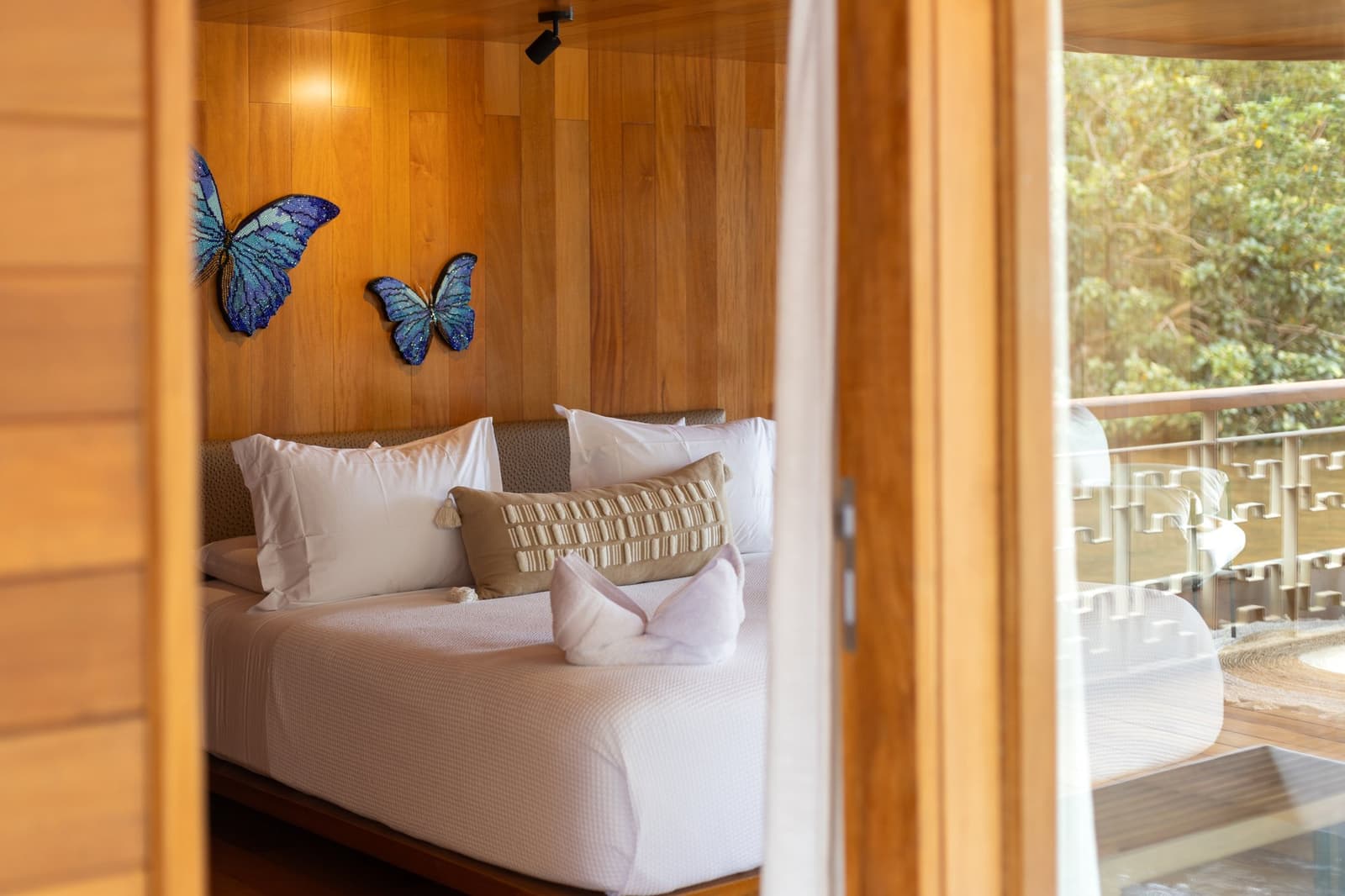 Borboletas morpho na parede, Amazônia na varanda. Essa noite vai ser boa. Quarto de suíte iluminado a quente no cruzeiro Delfin pelo Amazonas, fotografado através de portas de madeira abertas, com duas esculturas de parede de borboletas morpho azuis acima de uma cama de casal impecavelmente arrumada e uma varanda privativa com vista para a copa verde da floresta