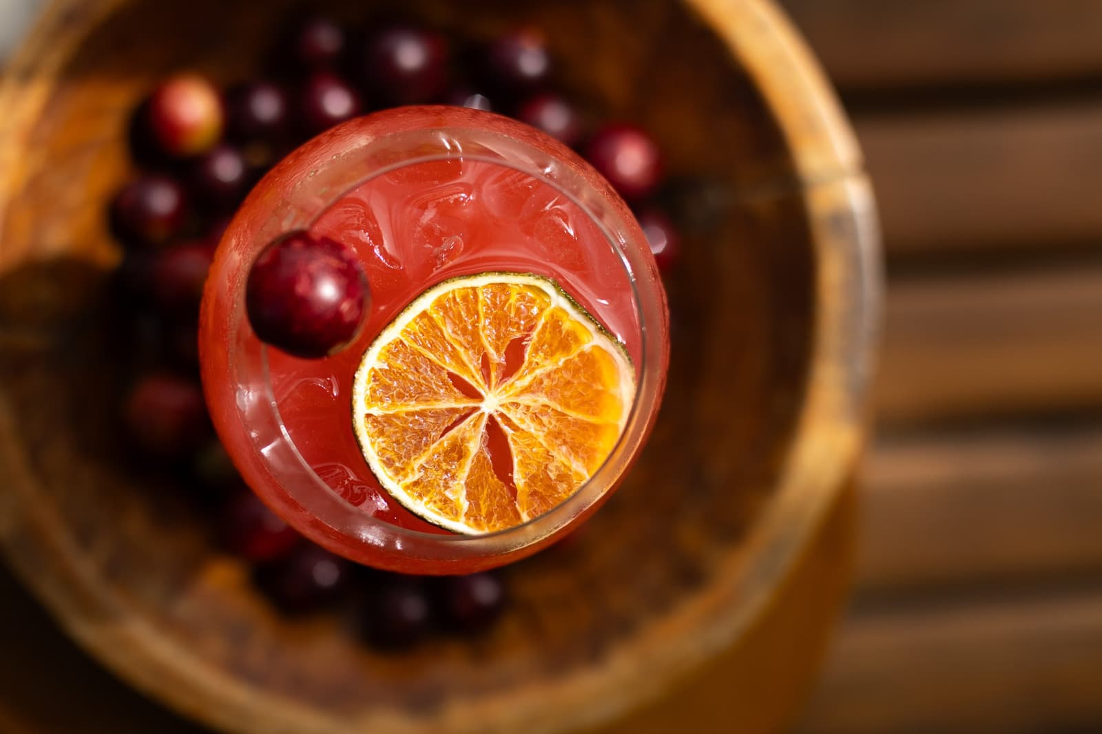 Capricho na taça, rio lá fora. Difícil não sorrir. Vista de cima de um drinque vermelho decorado com uma rodela de cítrico desidratado e um cranberry fresco, apoiado em uma tigela de madeira cheia de frutinhas escuras, servido no cruzeiro Delfin pela Amazônia