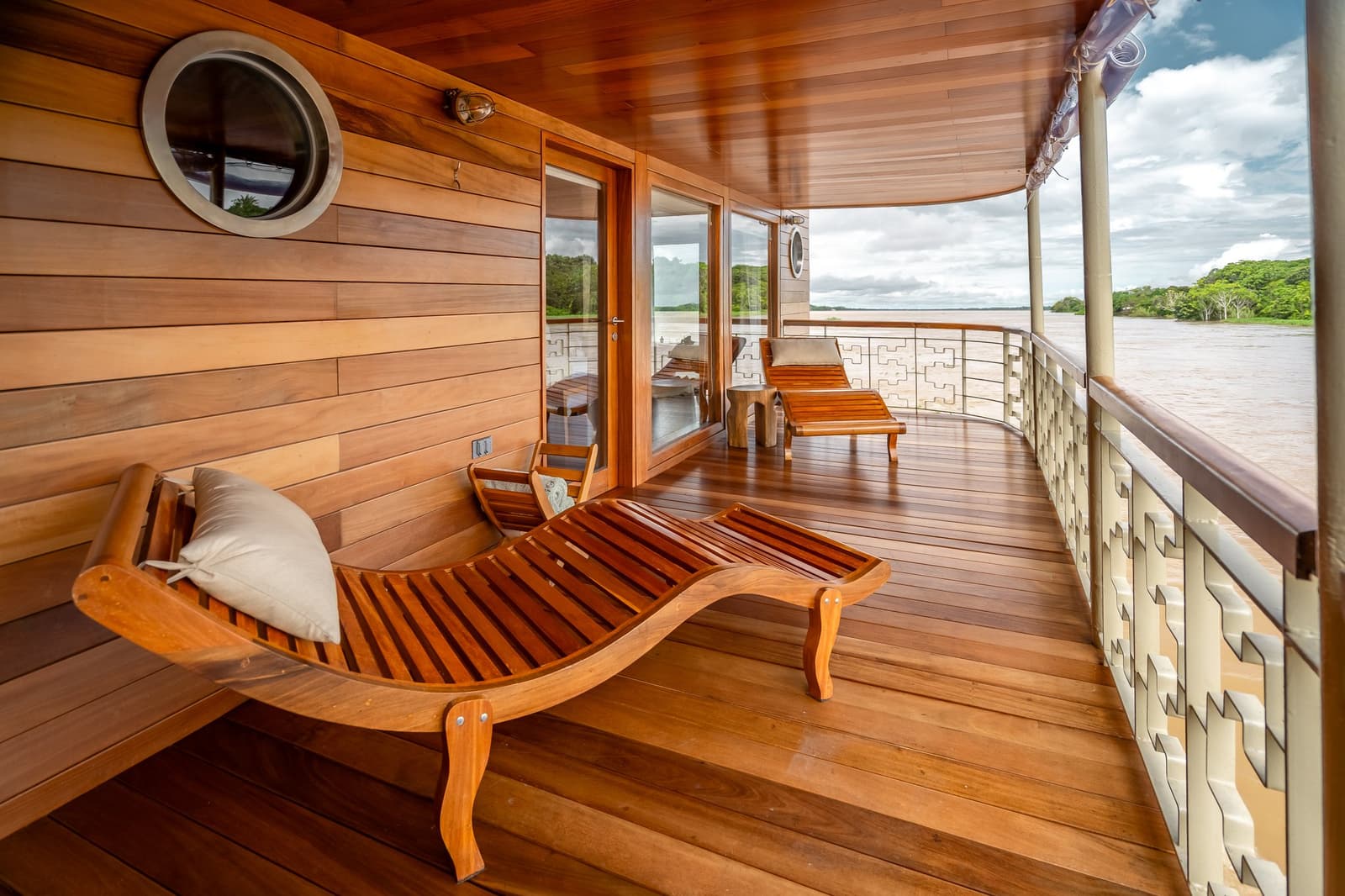 O rio passa, o tempo fica, aproveite cada detalhe. Photo: Mario Gomi Espreguiçadeiras de madeira curva no deck externo privativo de um navio de cruzeiro Delfin na Amazônia, com o amplo rio e a floresta verde visíveis além do corrimão ornamentado