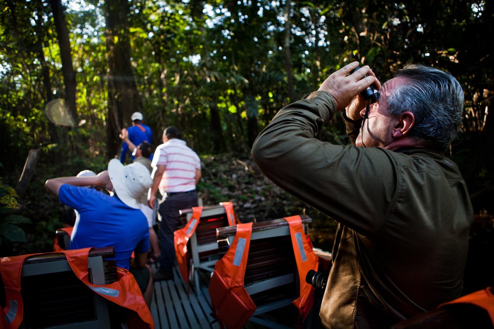 Todo mundo viu ao mesmo tempo, mas ele viu com mais detalhes. Photo: er: Rodrigo Rodrich Um homem levanta binóculos para observar a copa da floresta amazônica a partir de um pequeno skiff, enquanto outros hóspedes se inclinam para frente em direção à margem densamente arborizada