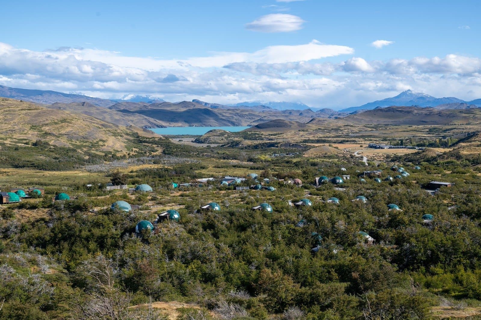 Domes in the scrub, a glacial lake beyond, your base is never boring. Aerial view of EcoCamp Patagonia showing dozens of green geodesic domes scattered among native shrubs, with a turquoise glacial lake and snow-capped Andean peaks visible in the background during puma trekking Patagonia expeditions