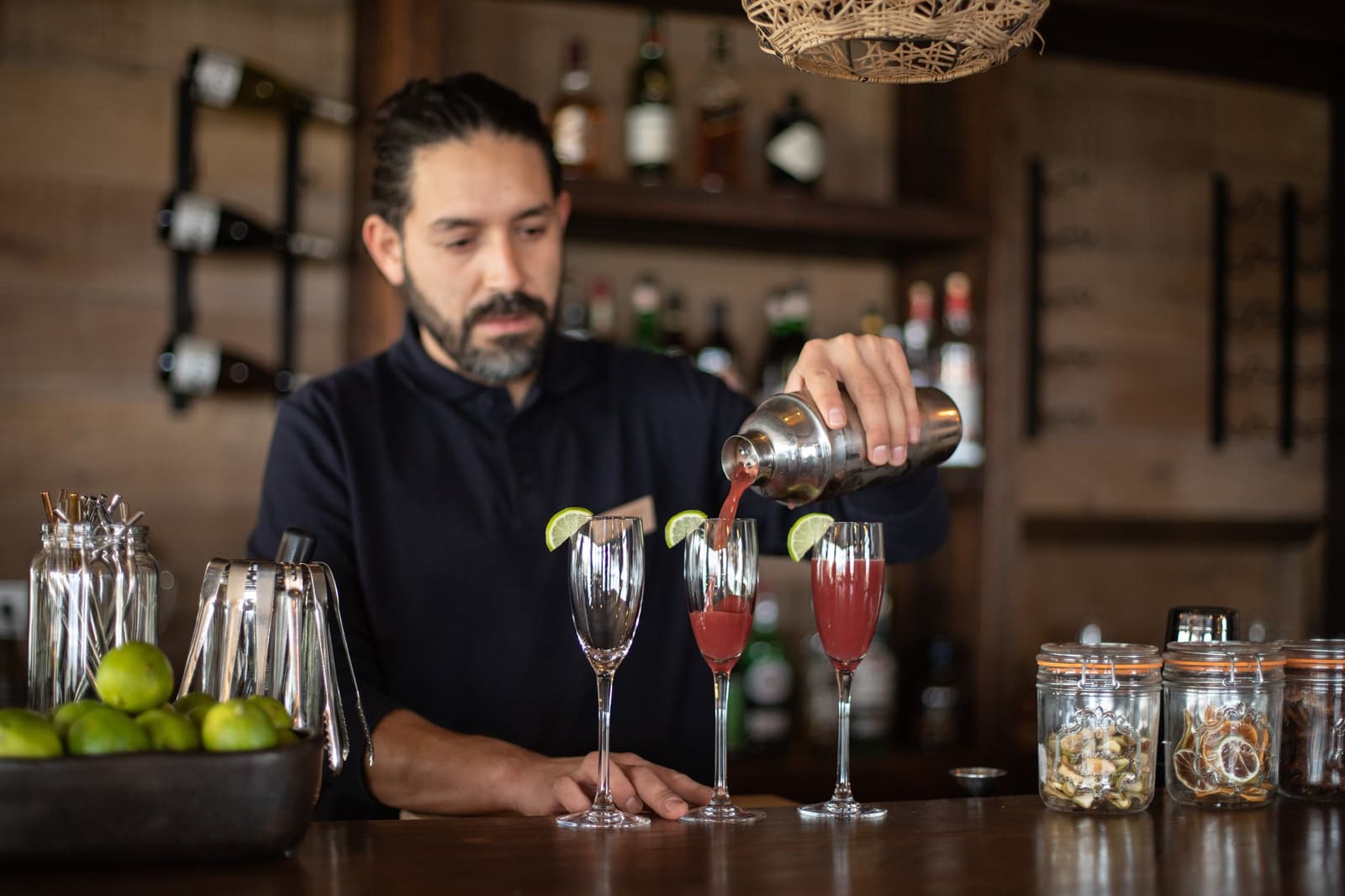 The open bar at the estancia takes its job seriously, lime always ready, shaker always cold. A bartender at Estancia Cerro Guido pours a deep-red cocktail from a shaker into tall flute glasses garnished with lime slices, behind a wooden bar stocked with spirits