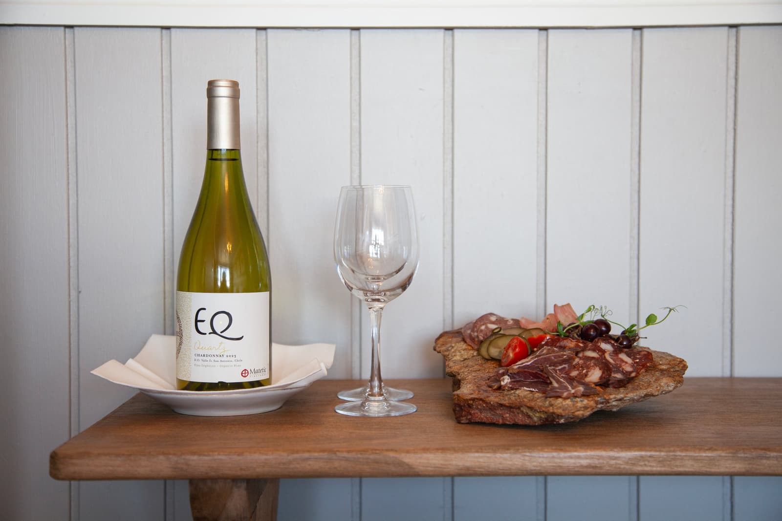 The estancia's idea of a warm hello: local charcuterie, a Chilean white, and no need to ask twice. A bottle of EQ Quartz Chardonnay 2023 by Matetic, a crystal wine glass, and a rustic stone board with cured meats, cherry tomatoes, pickles, and fresh herbs, arranged on a wooden shelf at Estancia Cerro Guido.