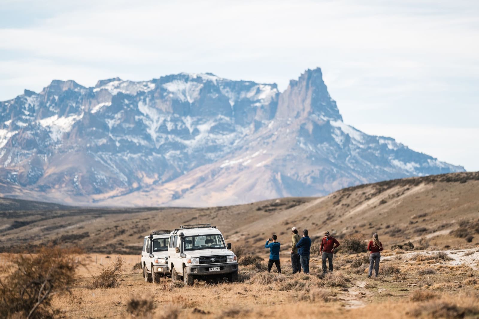 Pull over, everyone out. Some views earn a full stop. A group of five travelers stands beside a white Toyota Land Cruiser on the open Patagonian steppe during an estancia cerro guido excursion, with a dramatic snow-dusted granite peak rising sharply against a pale sky in the background.