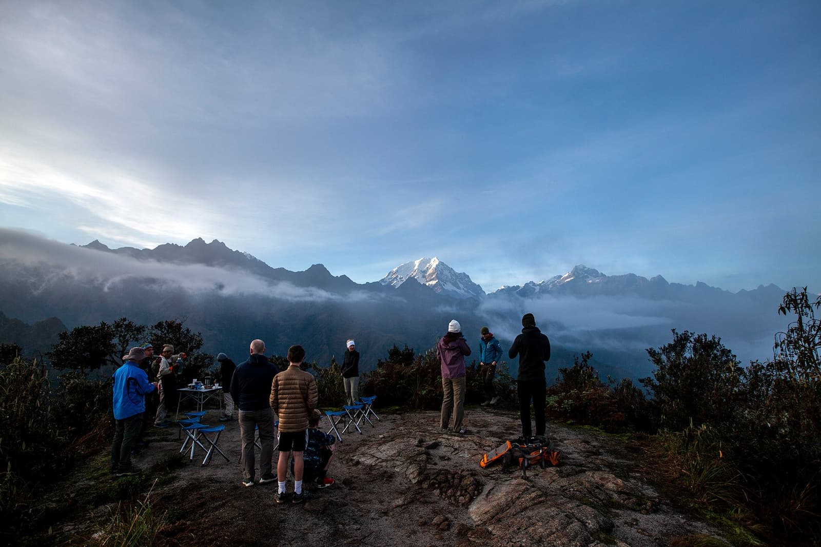 Tea in hand, mountains ahead, the porters had it sorted before anyone was fully awake. A small group of trekkers stands on a rocky high-altitude viewpoint at dawn on the Inca Trail to Machu Picchu, gazing at a snow-capped peak rising above a sea of cloud, with folding camp chairs and a small table set up behind them