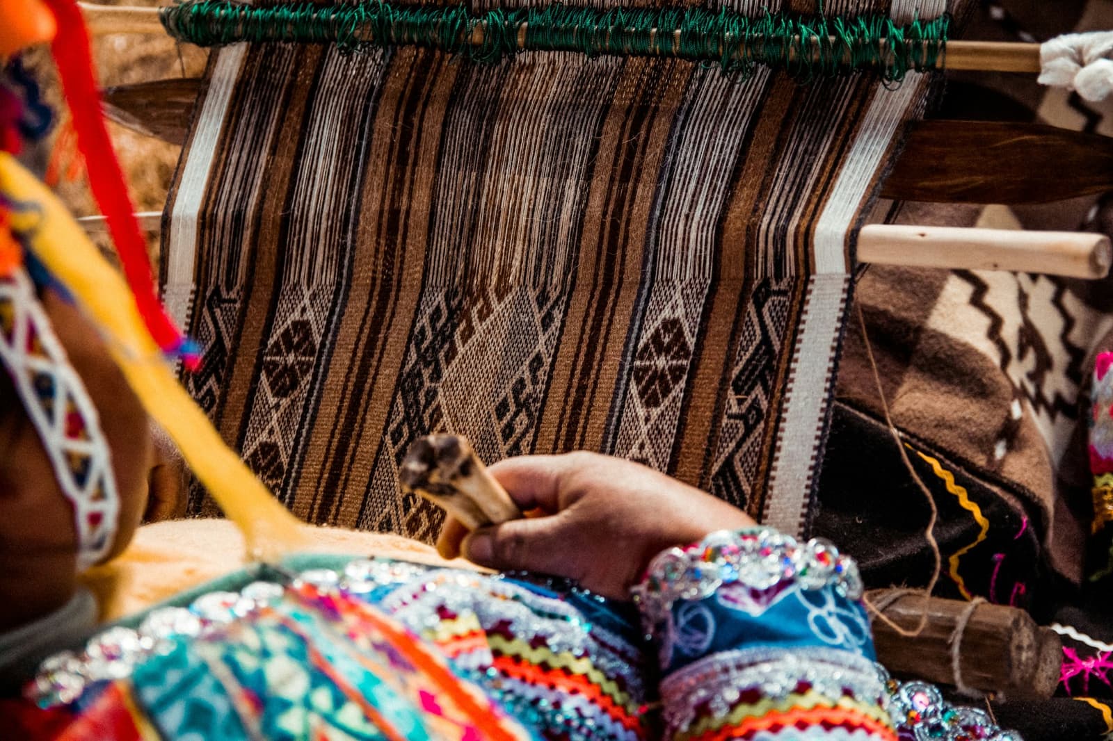 Every pattern carries a story that no guidebook has ever managed to translate. Close-up of a woman's hand guiding a bone shuttle across a traditional backstrap loom, weaving a brown-and-black geometric patterned textile, her sleeve decorated with colorful embroidery and beadwork, on this machu picchu amazon tour overland across Peru.