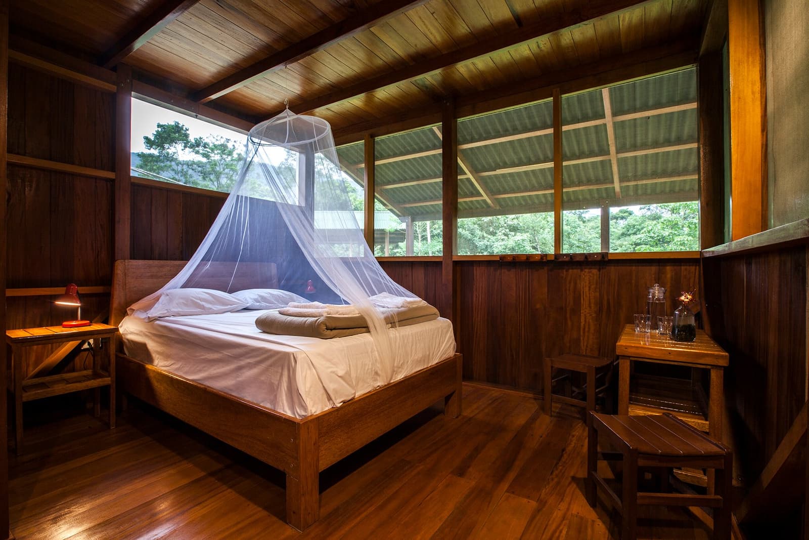 Sleep to the sound of the river, wake up to the forest. Rustic wooden cabin bedroom on a Machu Picchu and Amazon tour, featuring a double bed draped with a white mosquito net canopy, warm hardwood floors, a bedside lamp, and open louvred windows overlooking dense green forest canopy