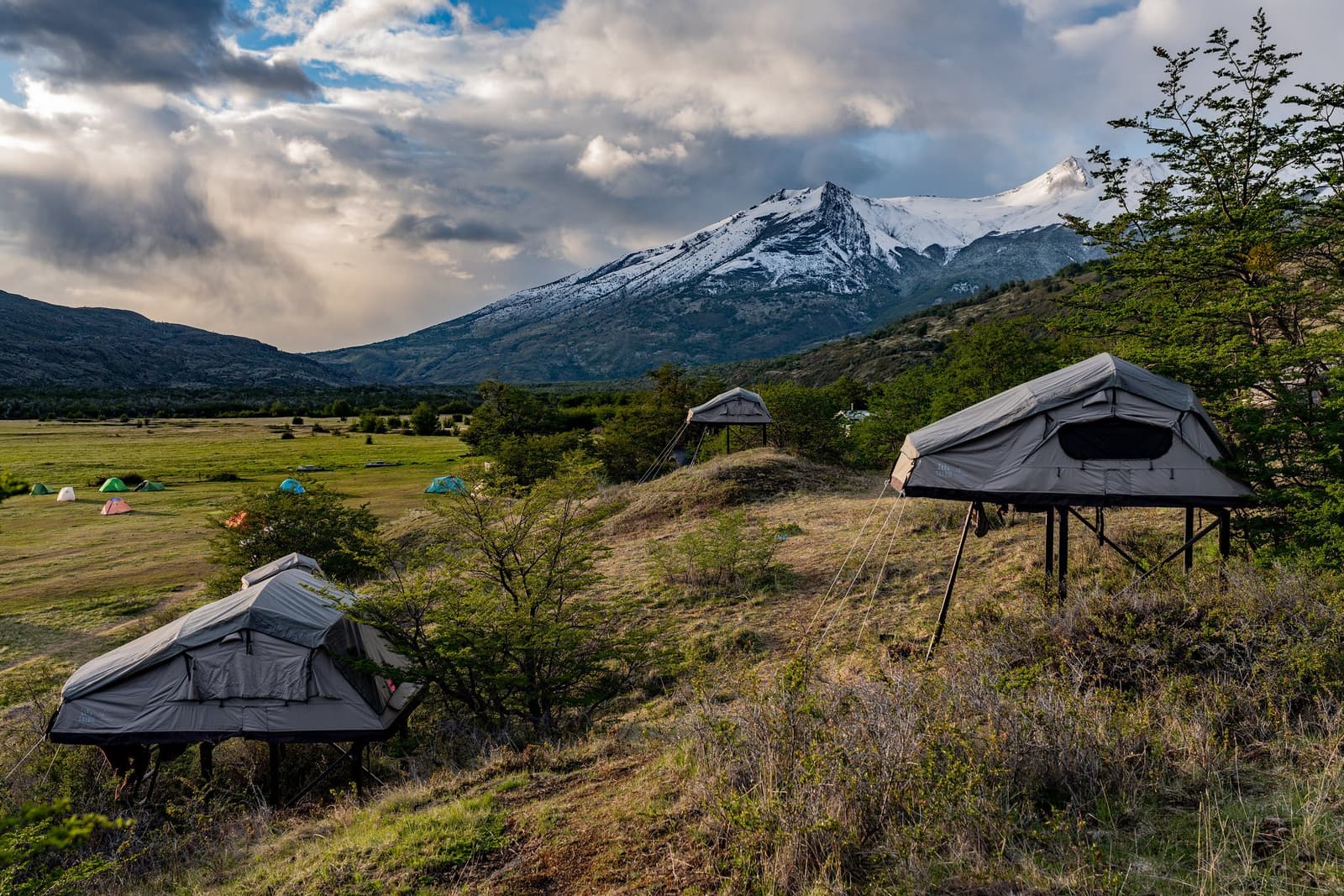 Sleeping off the ground, with that mountain watching over you all night. Photo: barria arjel Three elevated grey platform tents on a hillside during the self-guided O Circuit, with a snow-capped peak, dramatic storm clouds, and a wide green valley dotted with colourful dome tents in the background