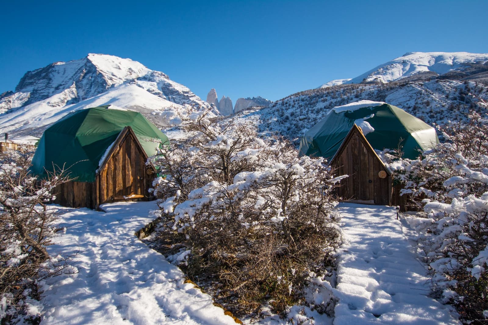Snow makes the domes look even more at home out here. Photo: Standard Dome Two green geodesic dome tents with wooden entrance panels dusted in fresh snow, set against snow-covered peaks and the distant granite spires of Torres del Paine on the o trek patagonia circuit