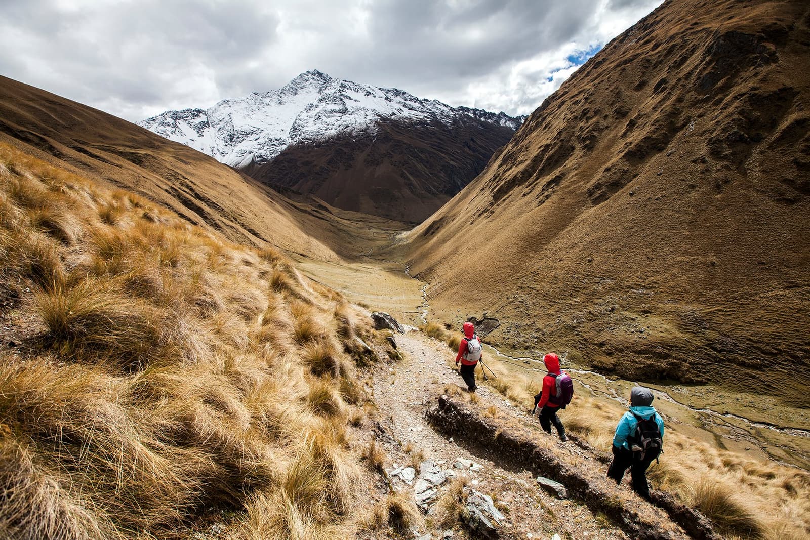 The valley is all yours, almost no one else comes this way. Three hikers with backpacks descend a narrow stone-edged trail on the off-the-beaten-path Salkantay trek, with golden puna grasslands flanking a wide glacial valley and a snow-capped peak rising under cloudy skies ahead