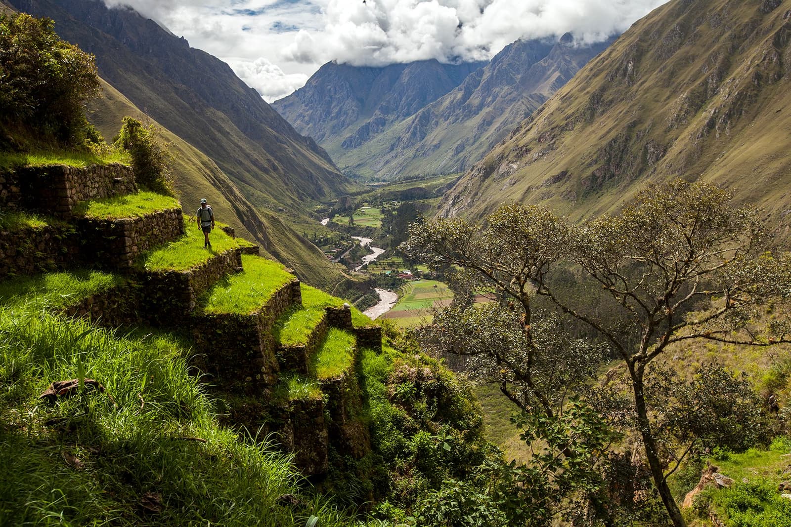 Terraces built for a civilization, now yours to walk alone. A lone hiker walks along ancient grass-covered Inca terraces on the off-the-beaten-path Salkantay trek, with a wide Andean valley, a winding river, and high mountain peaks stretching into the distance below