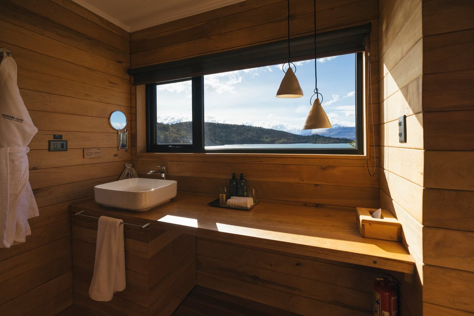 Brushing your teeth with that view feels like a small luxury you didn't know you needed. Photo: Patagonia Camp Warm cedar-clad bathroom at Patagonia Camp with a white vessel sink on a timber vanity, pendant lights, and a wide window framing Lake Toro and forested hills under a blue sky
