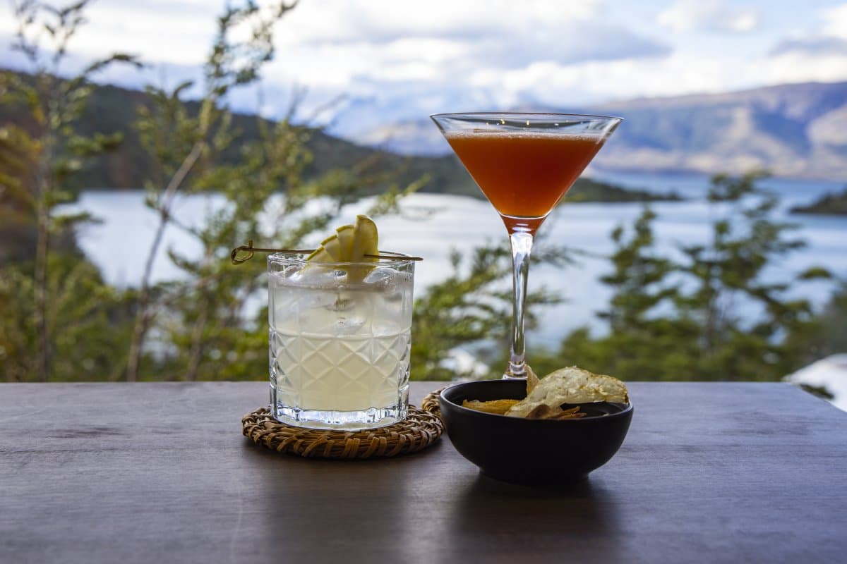 Two drinks, one view, and absolutely no reason to move. Photo: Patagonia Camp Two cocktails — a pale rocks drink garnished with apple and a deep amber martini — served with a bowl of chips on a wooden table at Patagonia Camp, with a lake and forested mountain range visible through the trees in the background