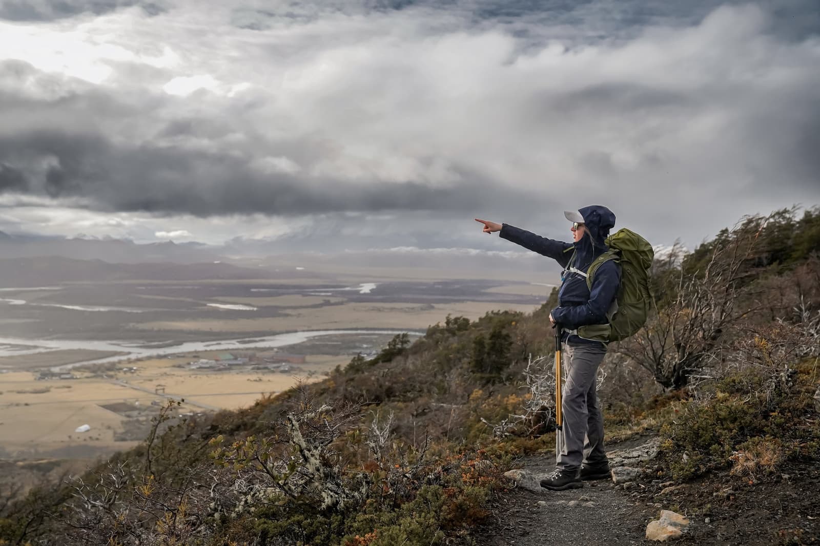 Someone always knows exactly what they're looking at out there. Photo: Patagonia Camp A hiker with a large green backpack and trekking pole stands on a mountain trail at Patagonia Camp, pointing toward a wide river valley below under a dramatic cloudy sky