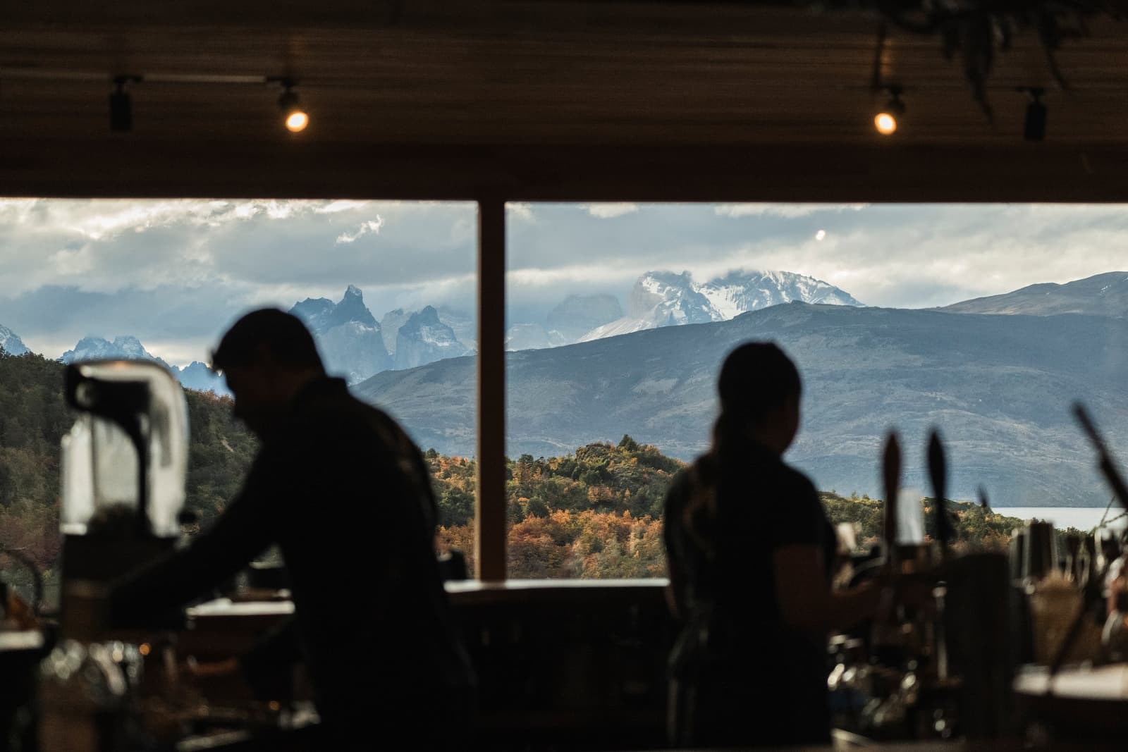 The kitchen crew, quietly keeping pace with one of the world's great views. Photo: Patagonia Camp Two silhouetted kitchen staff work at a restaurant counter at Patagonia camp, with large open windows framing snow-capped peaks and autumn-toned forest beyond