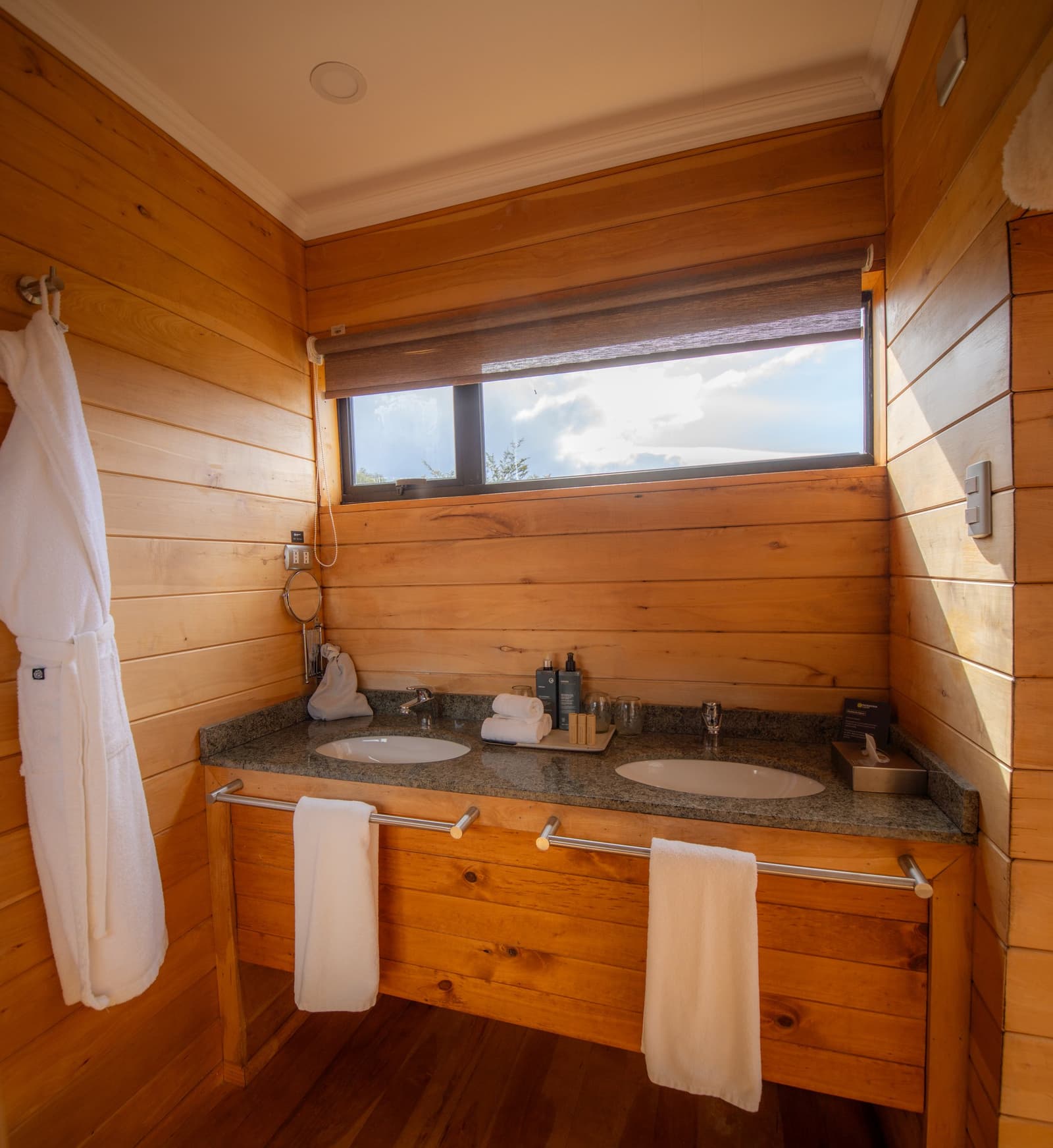 Two sinks, fluffy robes, and a sliver of Patagonian sky. Roughing it was never this comfortable. Photo: Patagonia Camp Double granite vanity with warm timber walls and white robes in a patagonia camp ensuite bathroom, with a narrow window opening to a sky view