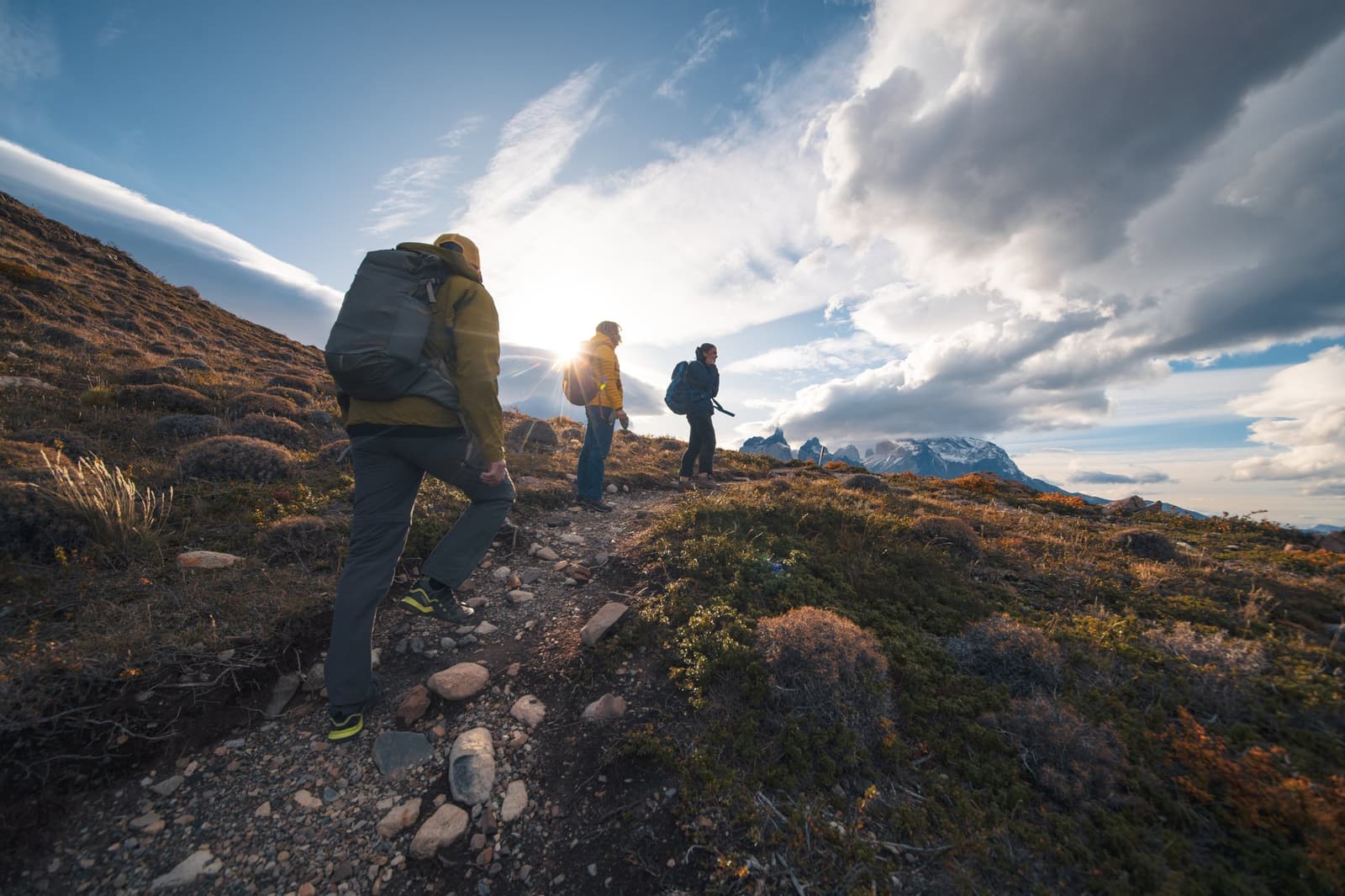Good boots, good company, and the Cuernos right there waiting for you. Photo: Patagonia Camp Three hikers with backpacks walk a rocky trail at golden hour on the Miradores route, with the granite spires of the Cuernos del Paine visible on the horizon under a dramatic sky during a patagonia camp excursion