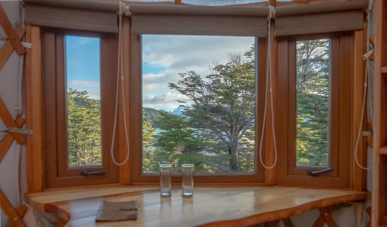 The view from your table is doing most of the work before breakfast even arrives. Photo: Patagonia Camp Curved wooden bay window of a patagonia camp yurt with two water glasses on a timber ledge and native lenga trees framing a glimpse of turquoise lake and mountain peaks beyond