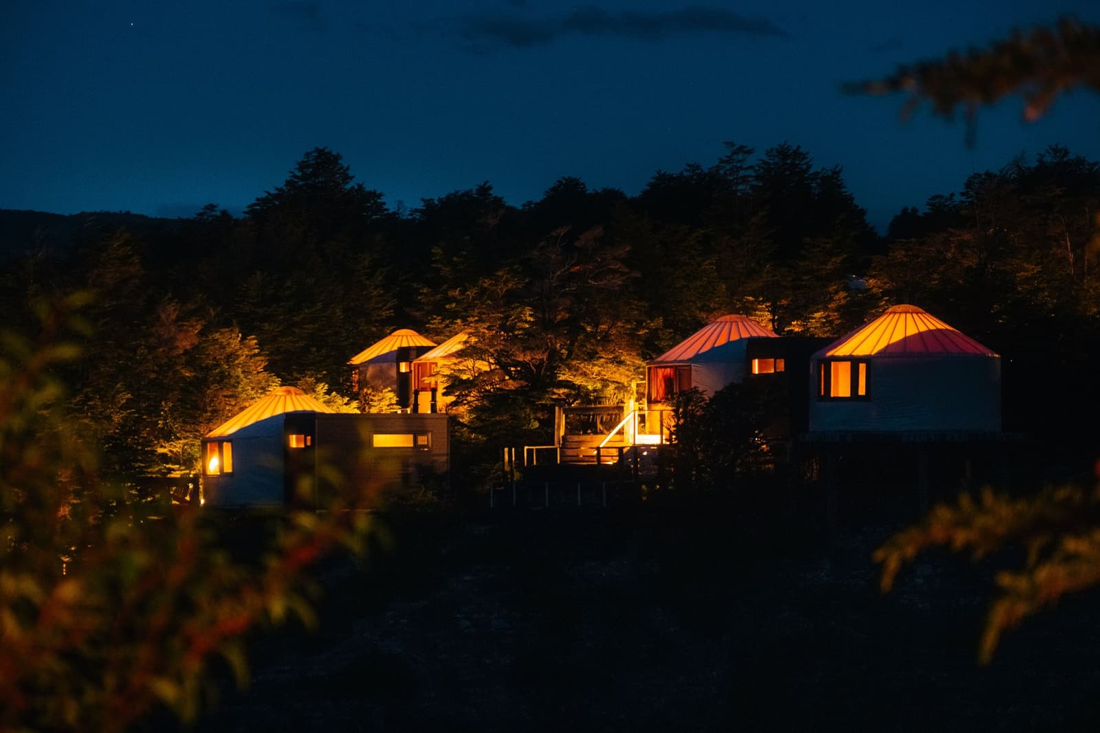 From out here in the dark, every lit window looks like a good reason to head back inside. Photo: Patagonia Camp Three illuminated patagonia camp yurts with warm orange rooftops glow against a deep blue twilight sky, nestled among native lenga beech trees above the lake shore