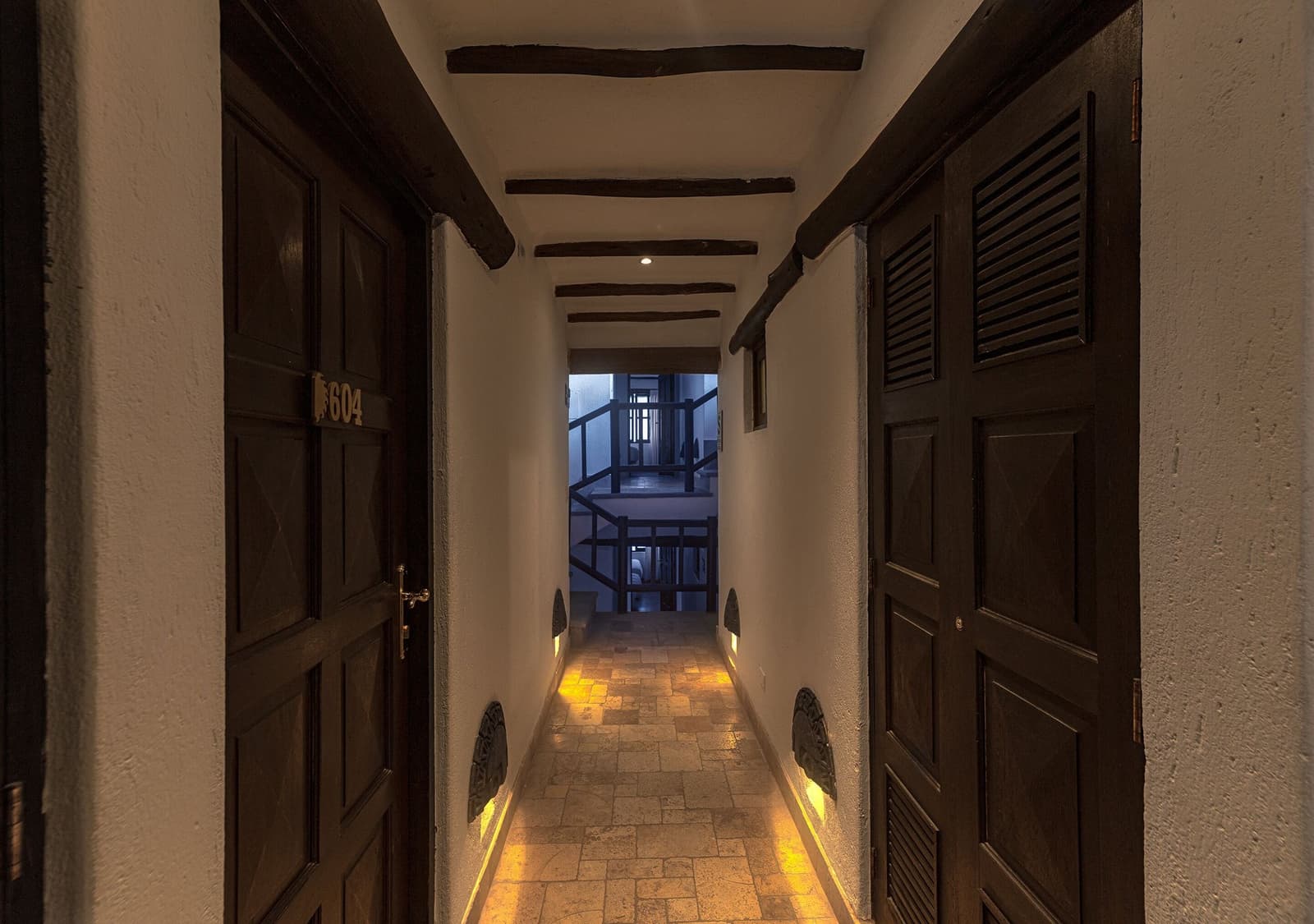 Vigas antigas, luz quente no chão, e uma cama esperando por você atrás daquela porta. Photo: Lucas Potographer Corredor de hotel com pouca luz, vigas de madeira aparentes no teto, piso de terracota, iluminação âmbar no chão, portas de madeira escura incluindo o quarto 604 e uma escada visível ao fundo, proporcionando descanso confortável no trek Salkantay de 5 dias