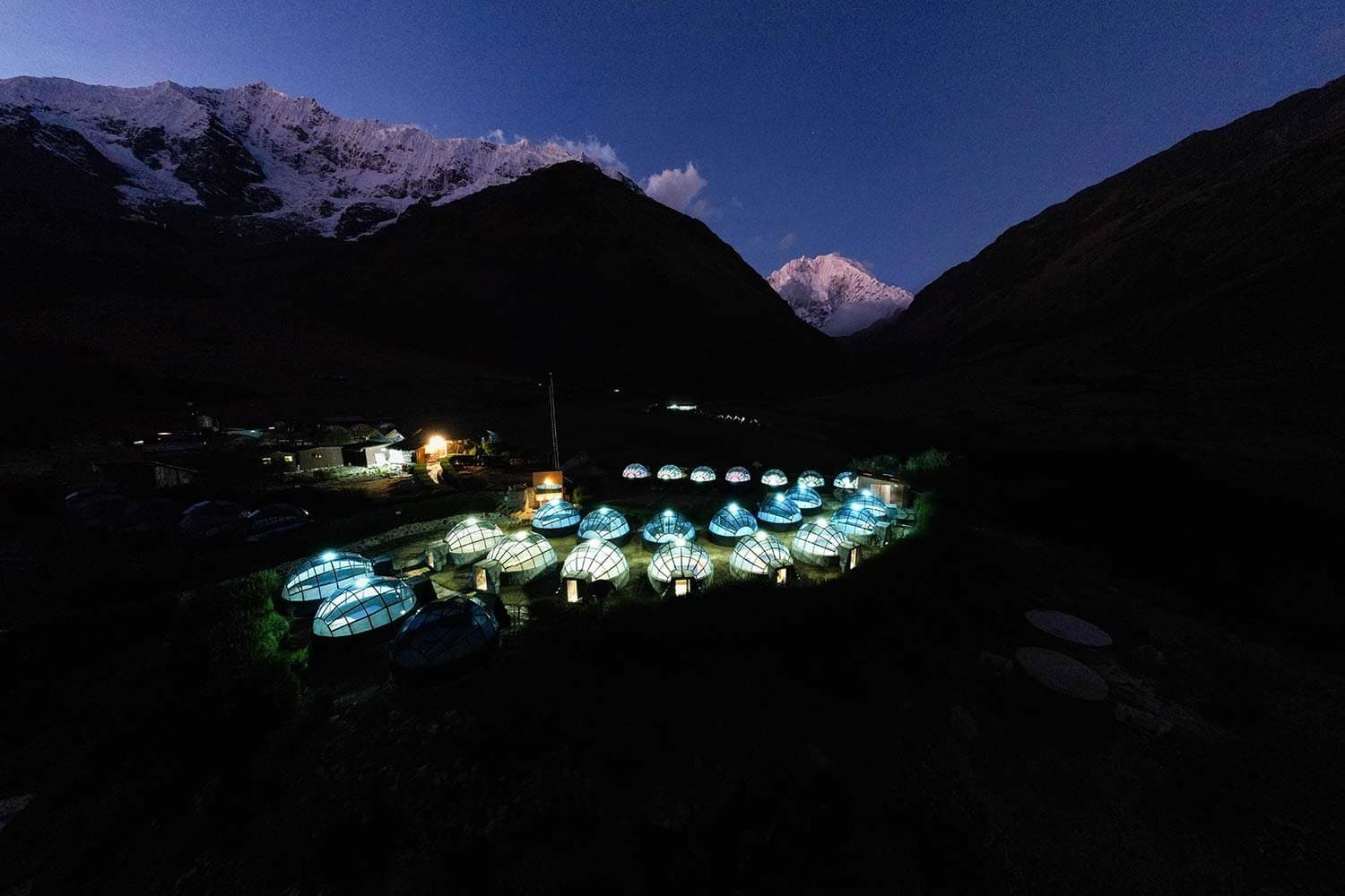Vinte domos acesos no vale, montanhas nevadas ao fundo, e uma cama te esperando lá dentro. Photo: Sky Camp Soraypampa Vista aérea noturna de domos geodésicos iluminados em um acampamento de altitude no trekking Salkantay de 5 dias, com picos nevados dos Andes brilhando sob um céu azul profundo ao entardecer
